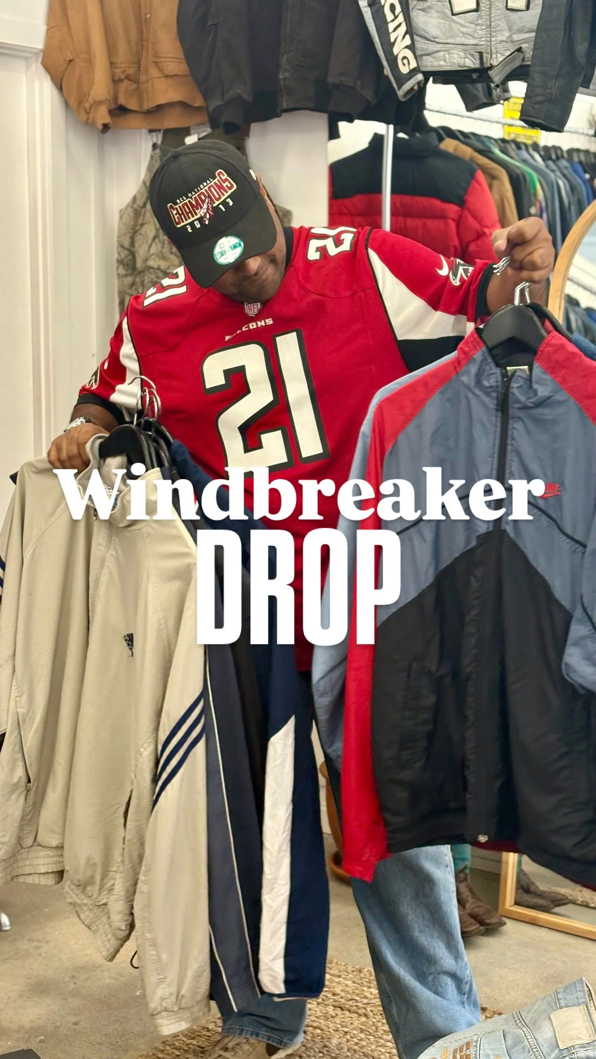 Person wearing a red Atlanta Falcons football jersey and a black Baseball cap, shopping for clothes in a store with jackets and pants hanging on racks.