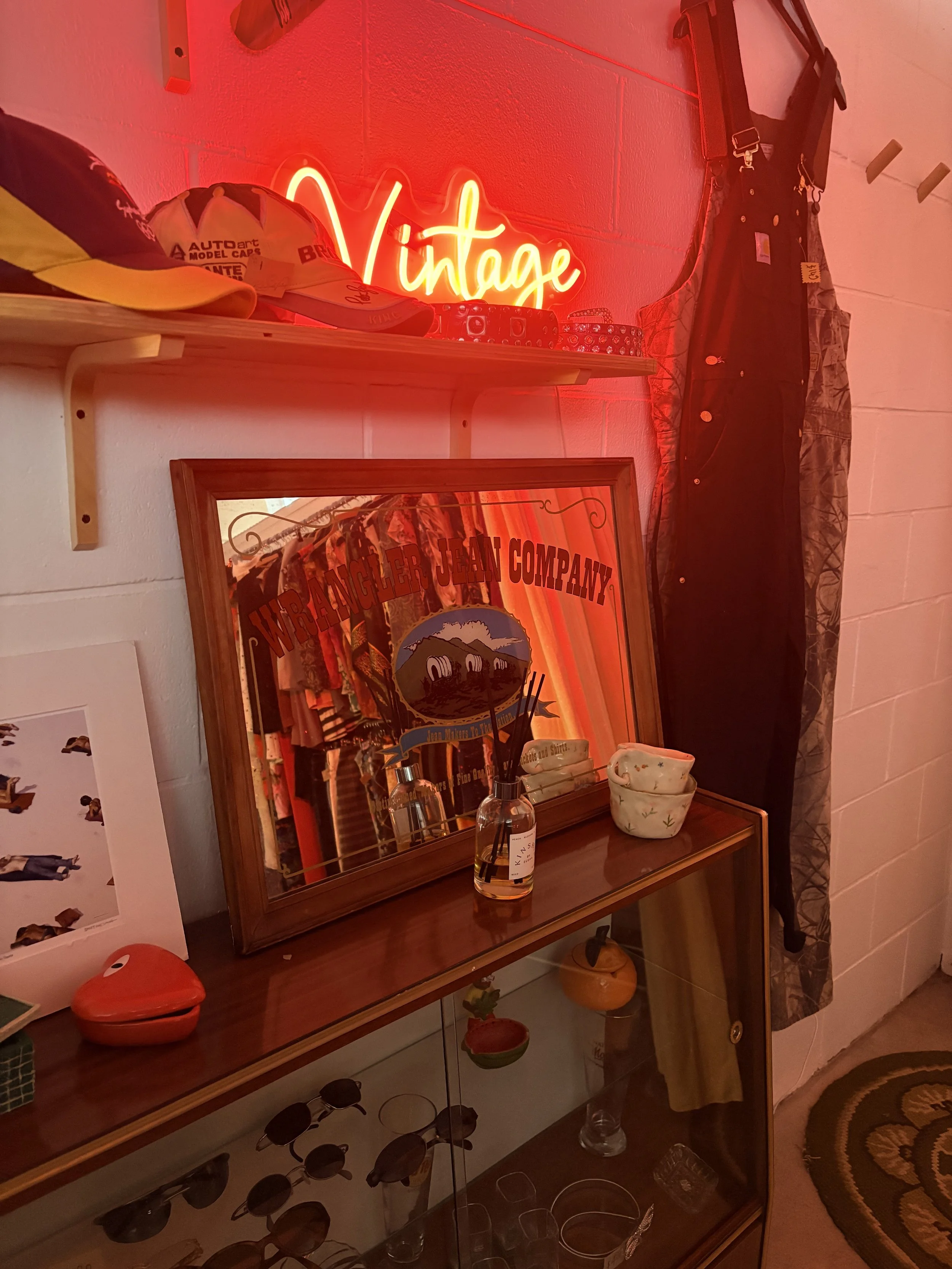 A wooden shelf with vintage items including a colorful cap, a neon 'Vintage' sign, a framed Wabler Jean Company poster, a perfume bottle, and ceramic bowls. There's a coat or curtain hanging beside the shelf, and a glass display cabinet with sunglasses and glasses underneath.