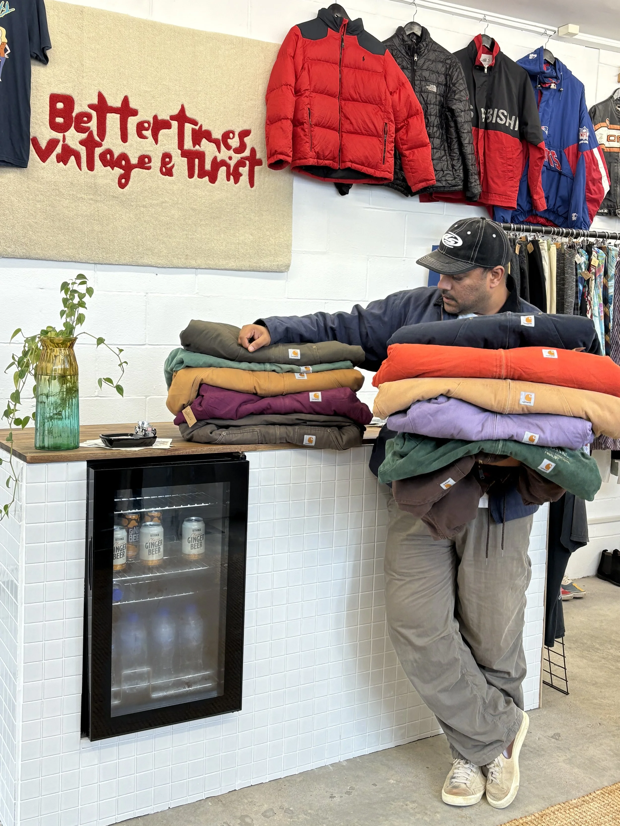 A man in gray pants, a black jacket, and a black cap is holding a stack of colorful clothes, including purple, yellow, red, green, and gray, while leaning against a white tiled counter with a glass door mini fridge underneath. The mini fridge contains a few cans of ginger beer. To the left, a green and yellow glass vase with a plant is on the counter. Behind him, there are jackets hanging on the wall and a beige wall with a red text sign that says "Better Thrift Vintage & Thrift."