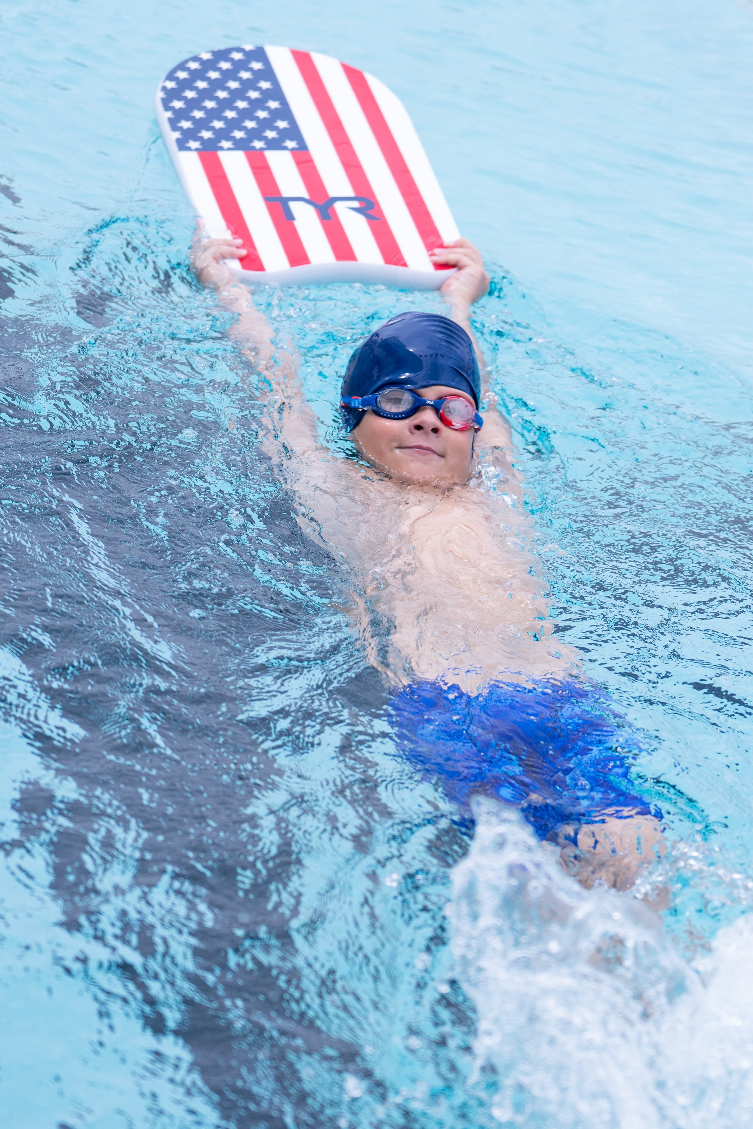 Child swimming in a pool with a patriotic American flag-themed floatboard, wearing swimming goggles and a swim cap, smiling at the camera.