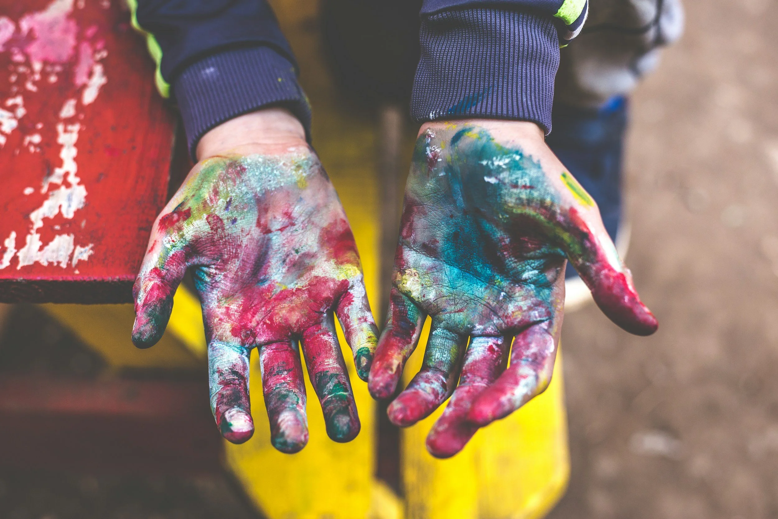 Two hands covered in colorful paint, with visible smudges and streaks, resting on a red bench, with a blurred background.