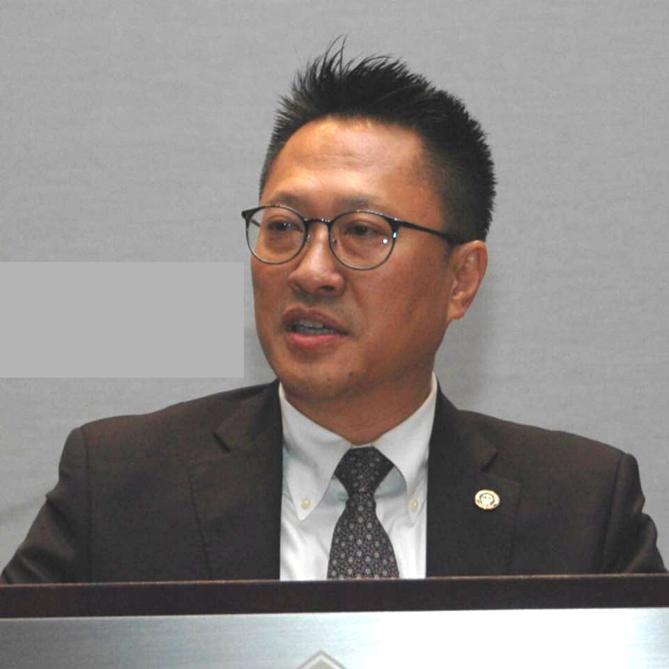 A man wearing glasses, a black suit, white shirt, and patterned tie, seated at a desk.