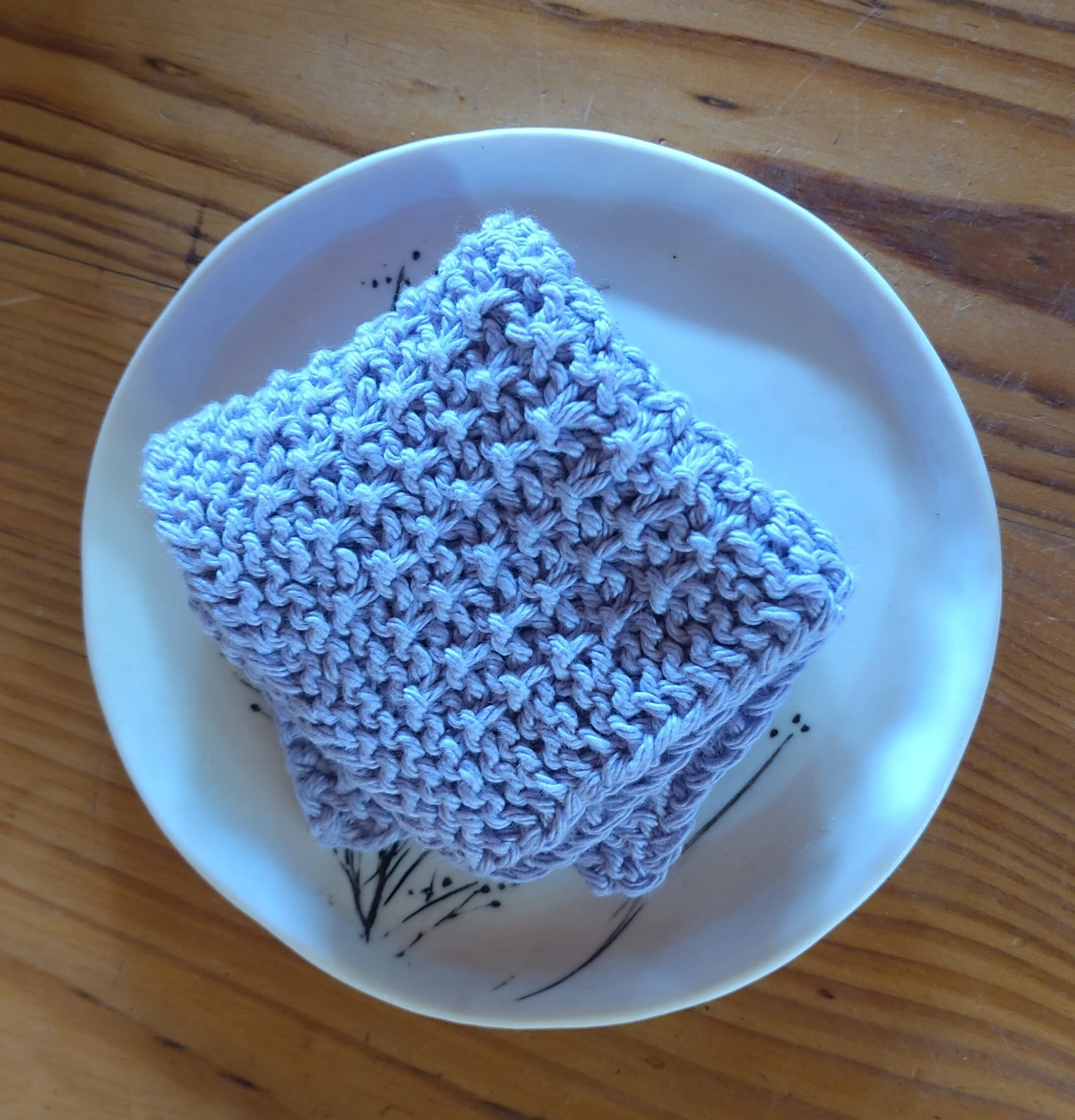 A knitted gray washcloth resting on a white ceramic plate with black line art design, placed on a wooden surface.