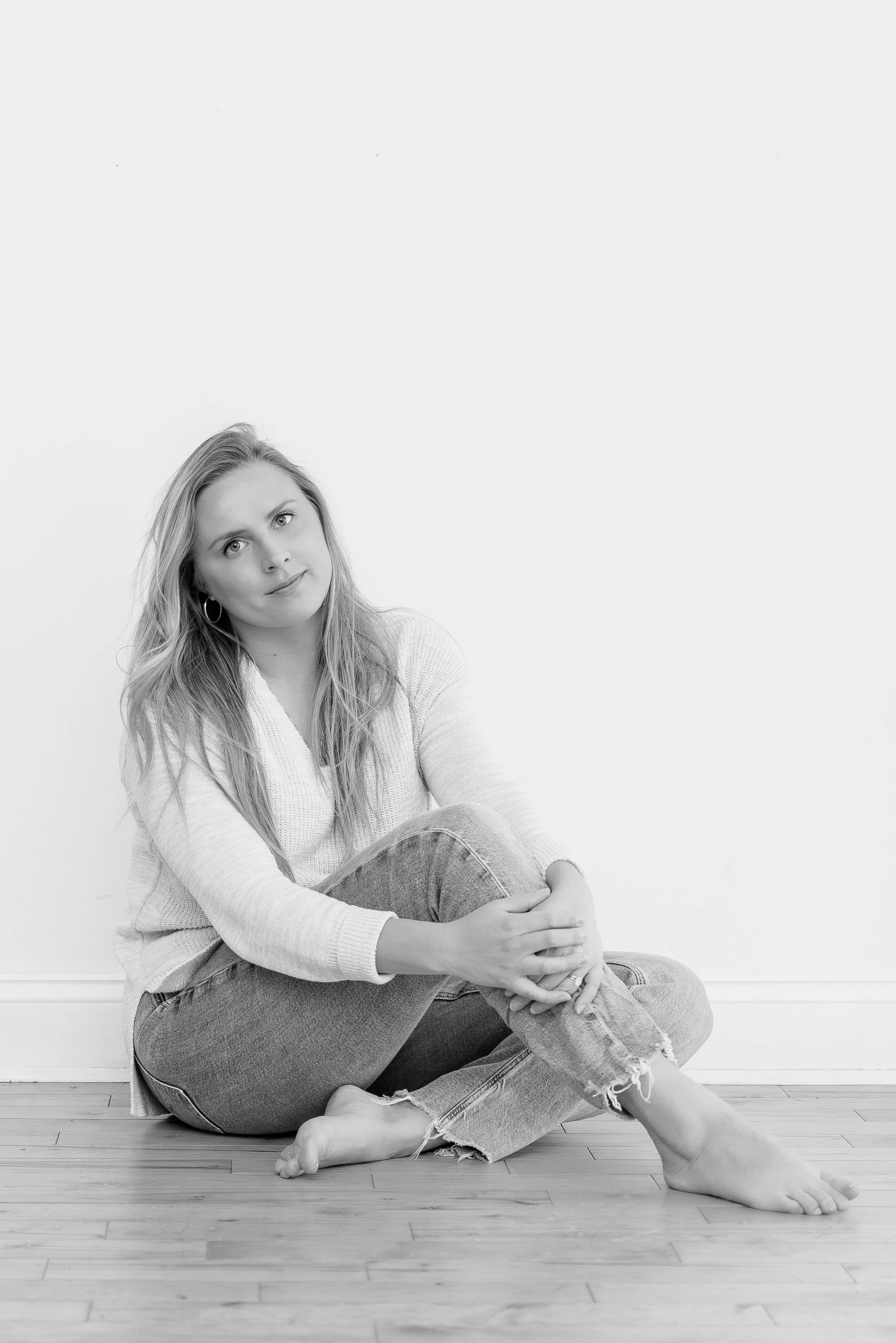 A black and white photo of a woman sitting on the wooden floor against a plain white wall, with her arms wrapped around her raised knee, looking at the camera.