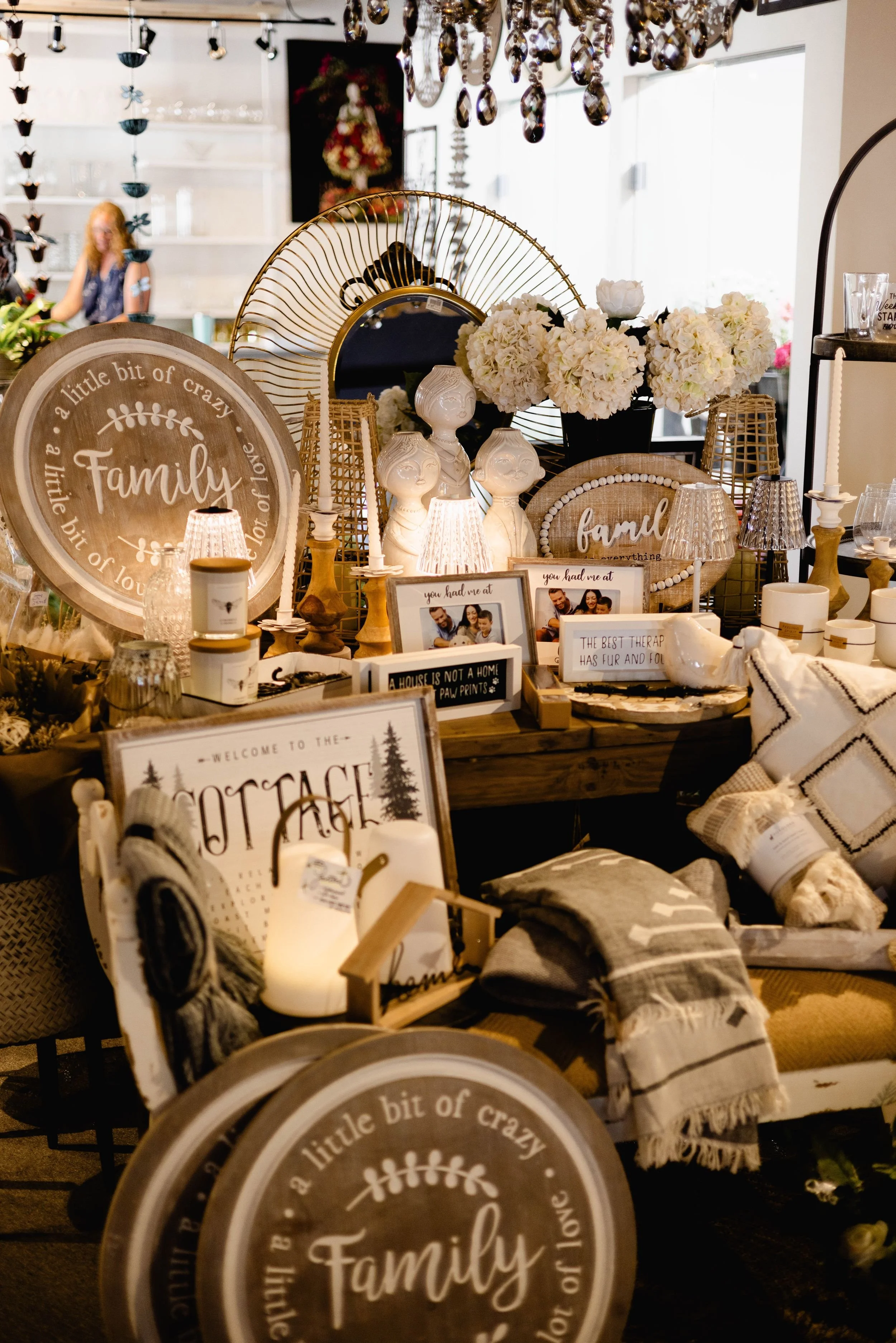 A display of home decor and gift items, featuring signs with messages about family, flowers, and cozy living, set against a well-lit background with reflections of a window and hanging chandeliers.