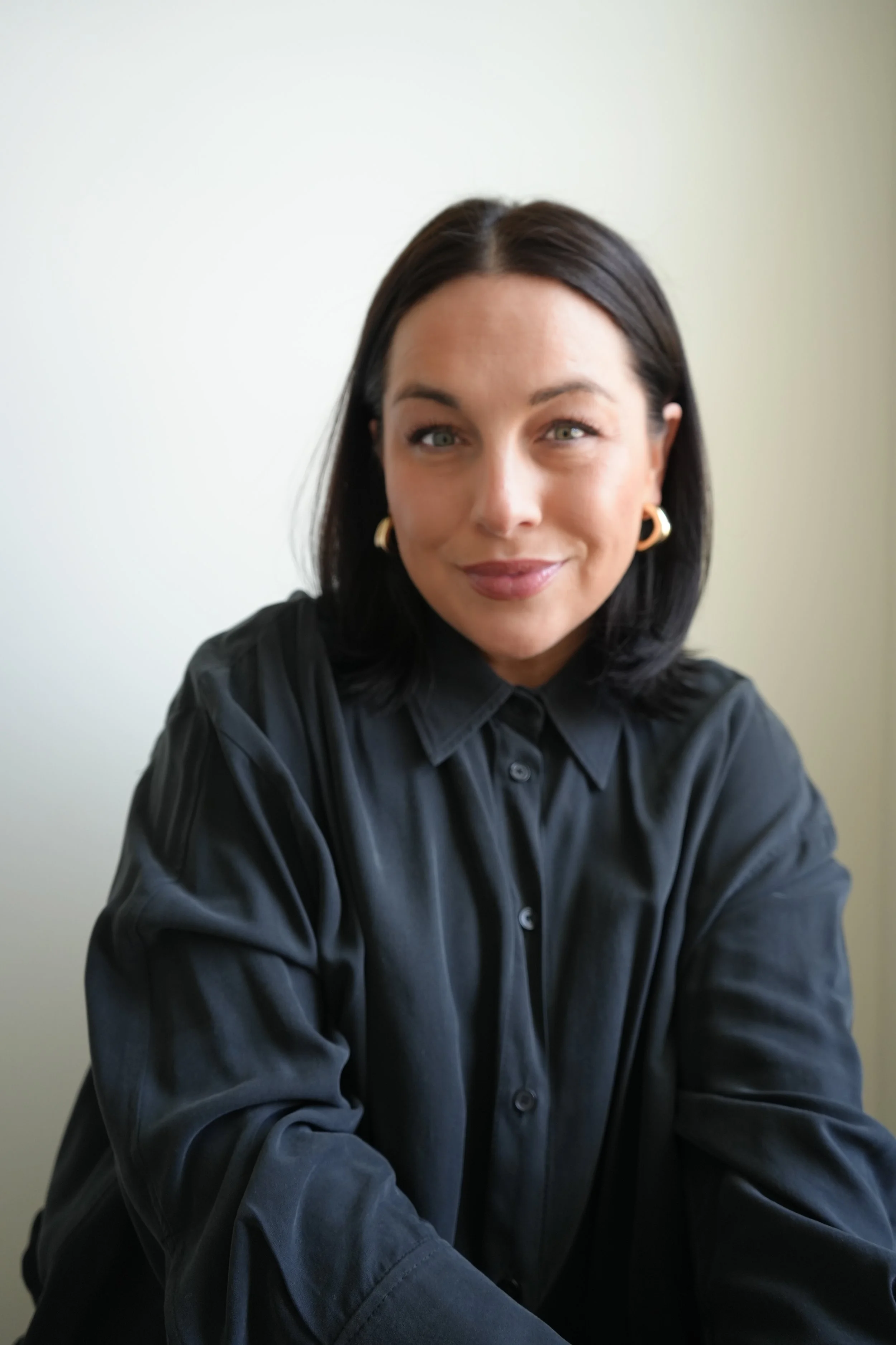 A woman with dark hair, wearing hoop earrings and a black shirt, smiling at the camera. She is sitting against a plain light-colored wall.