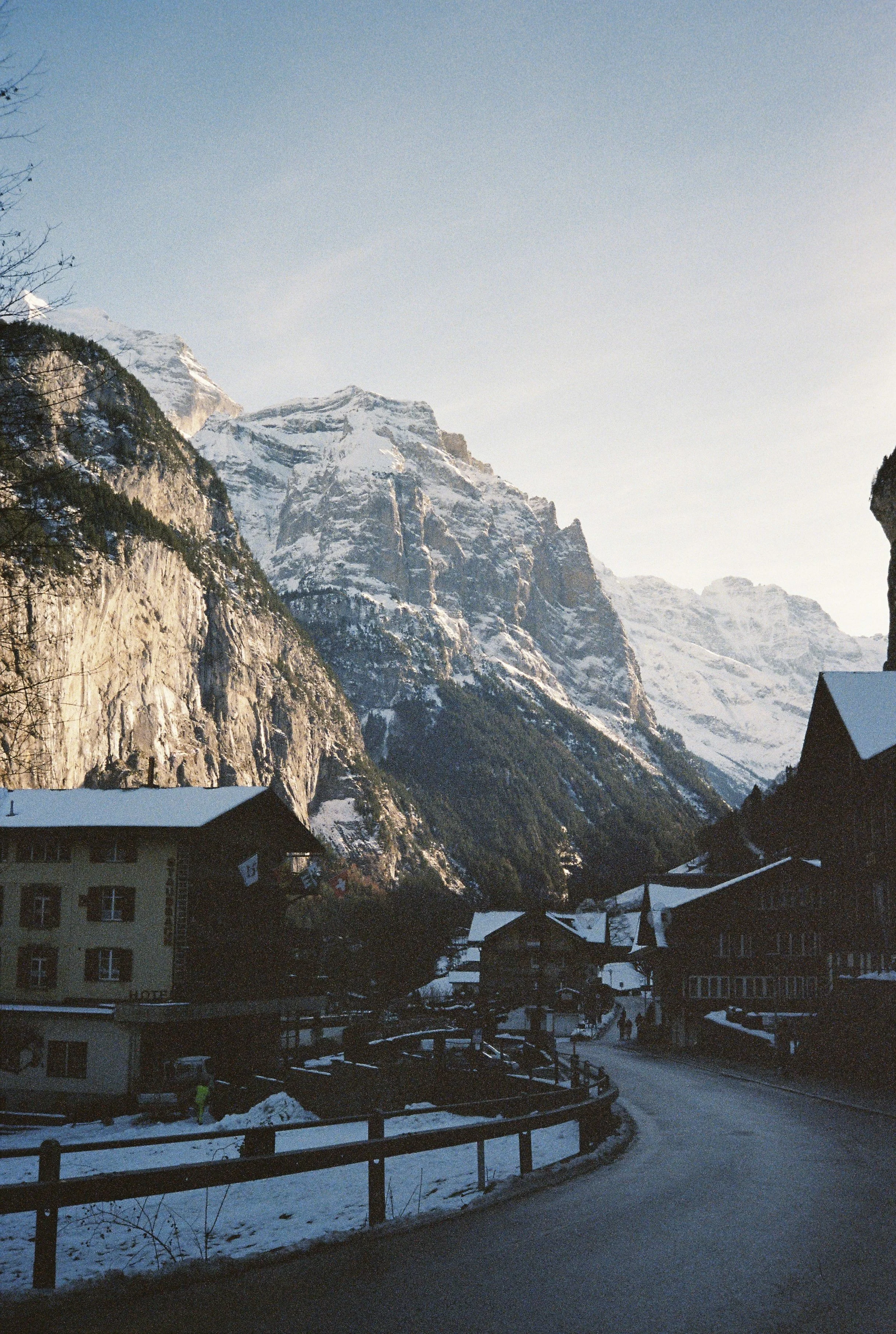 Snow-covered buildings and a road in a mountain town, with towering snow-capped mountains in the background, during daytime.