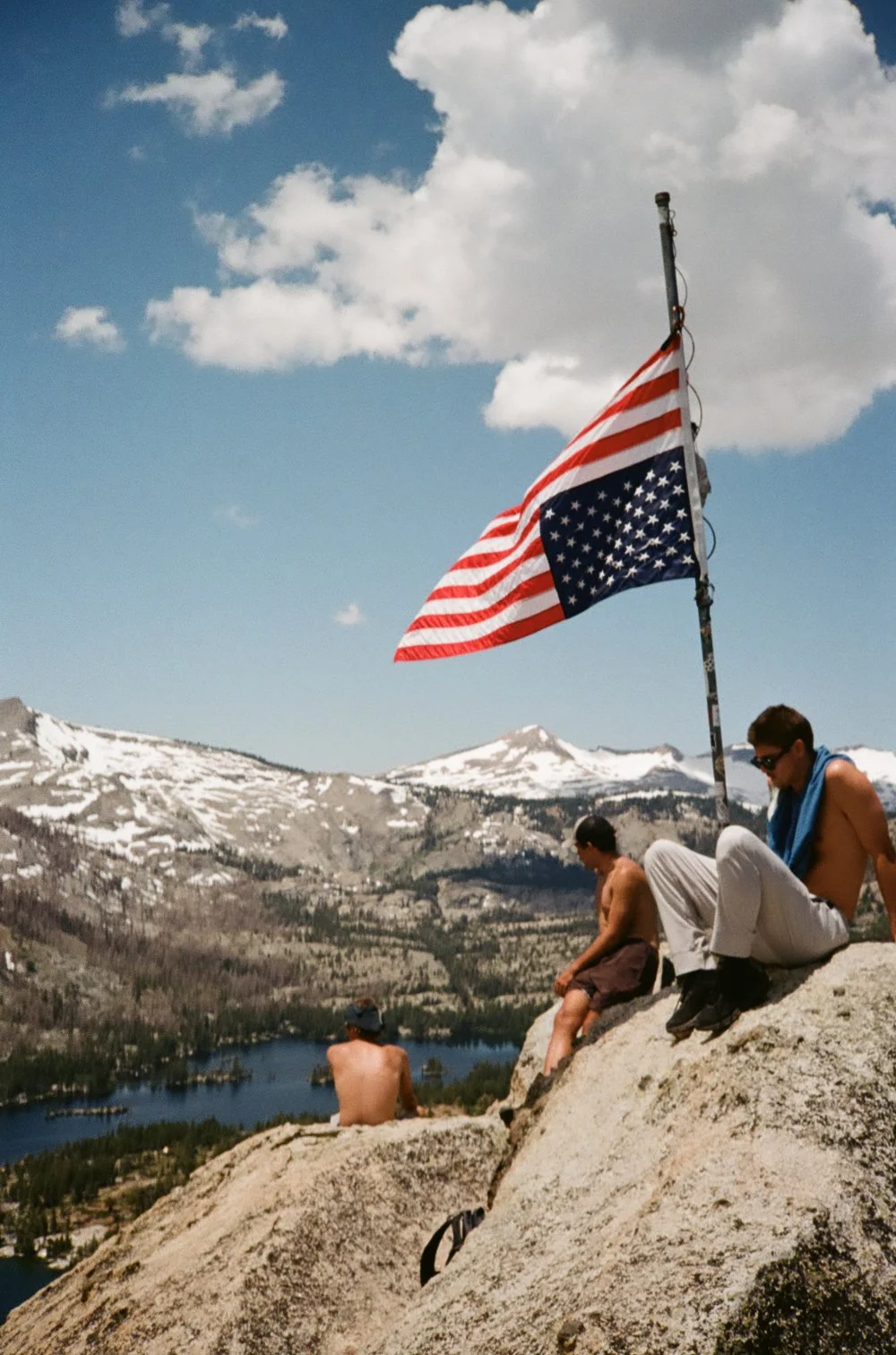 People sitting on rocky ledge with a tattered American flag on a pole, overlooking a mountainous landscape with snow-capped peaks, forested area, and a lake, under partly cloudy sky.