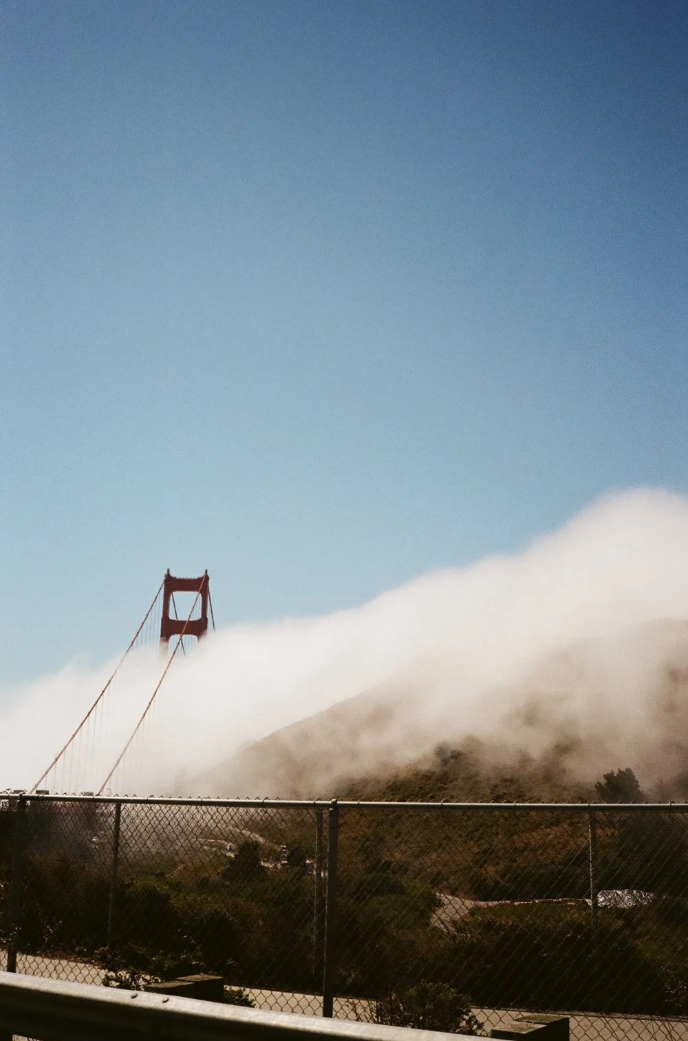 The Golden Gate Bridge partially obscured by fog, with a clear blue sky above and a chain-link fence in the foreground.