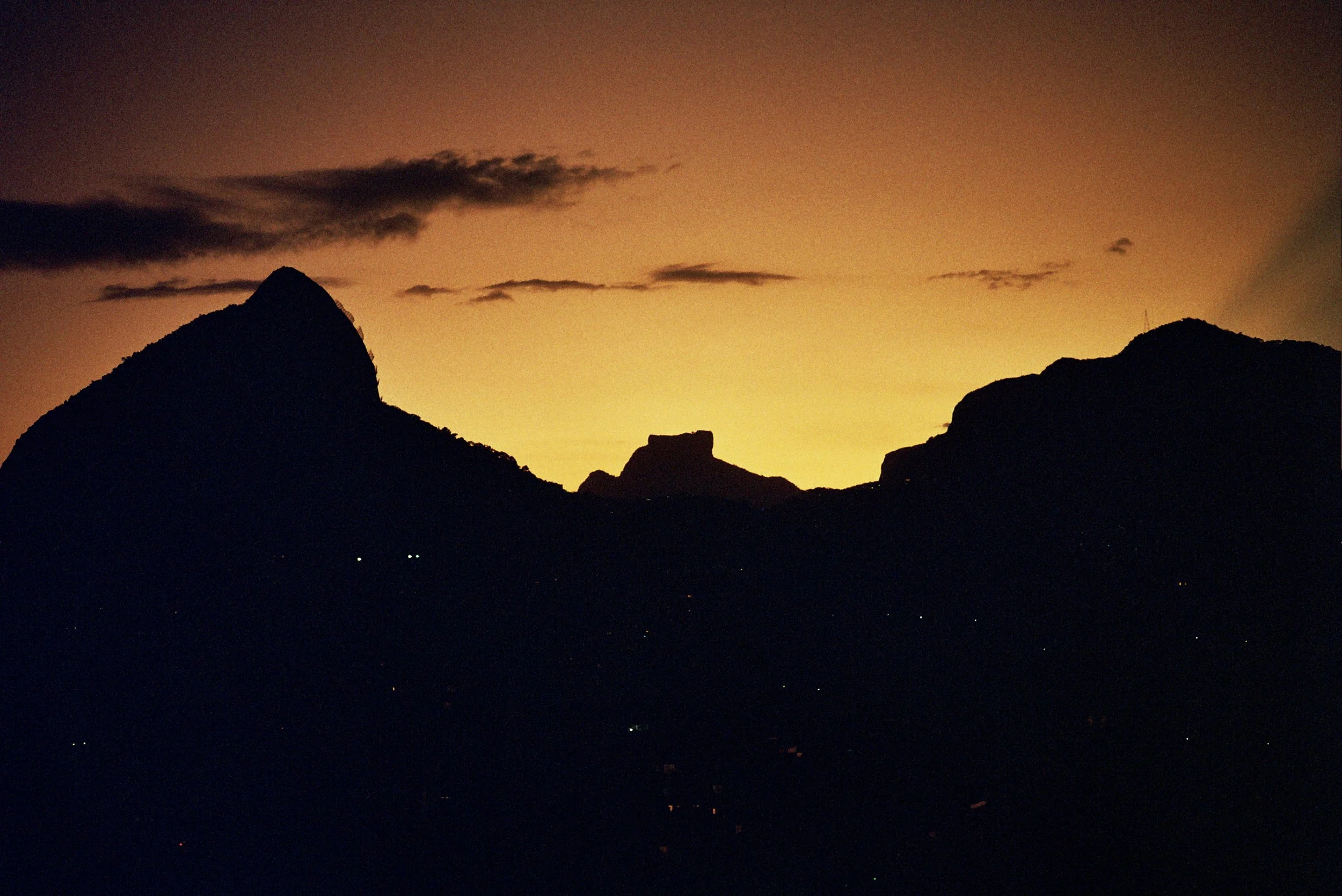 Silhouetted mountains against a sunset or sunrise sky with dark clouds and small lights at the base.