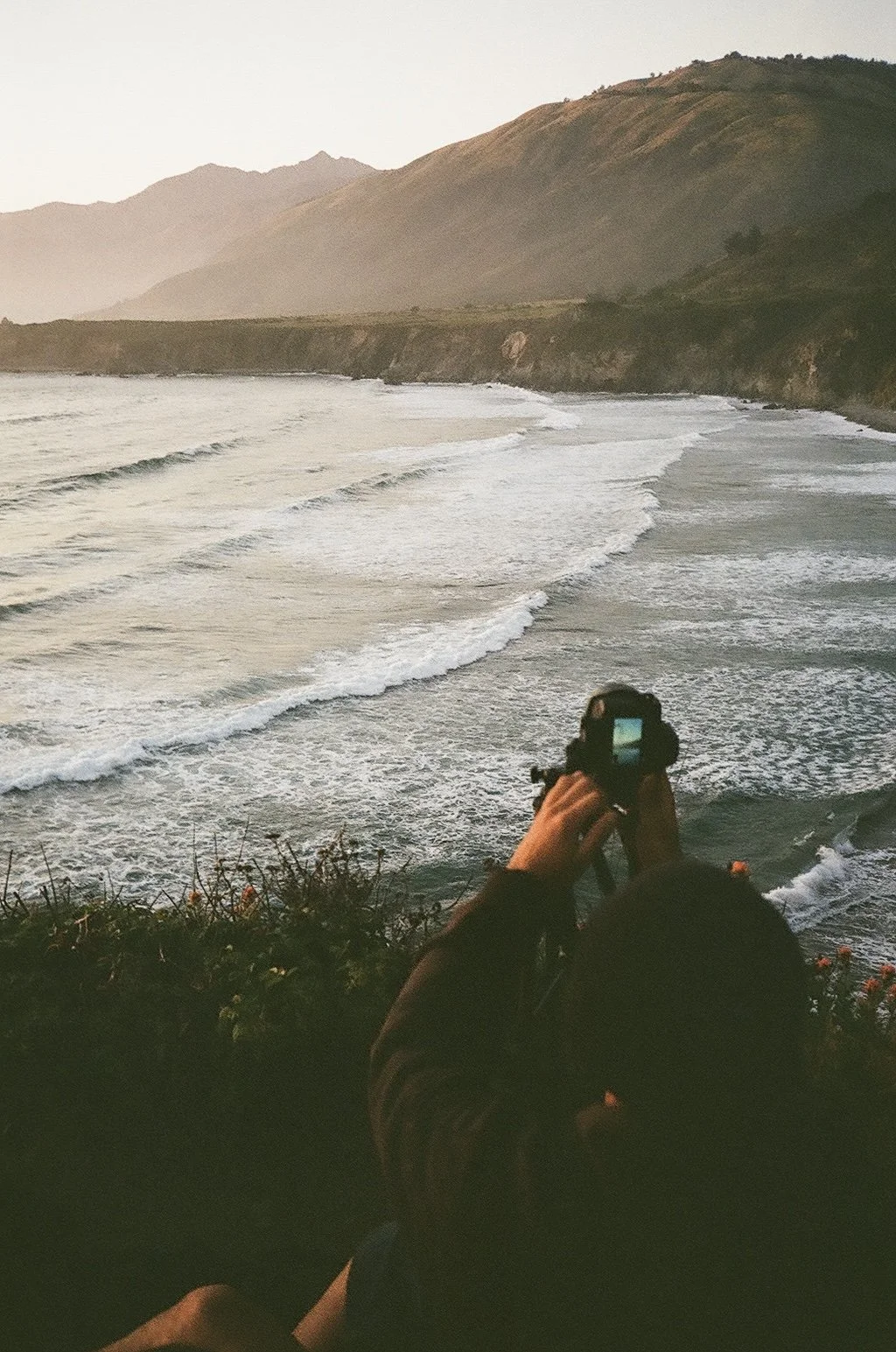 Person taking a photo of the ocean with waves and mountains in the background during sunset.