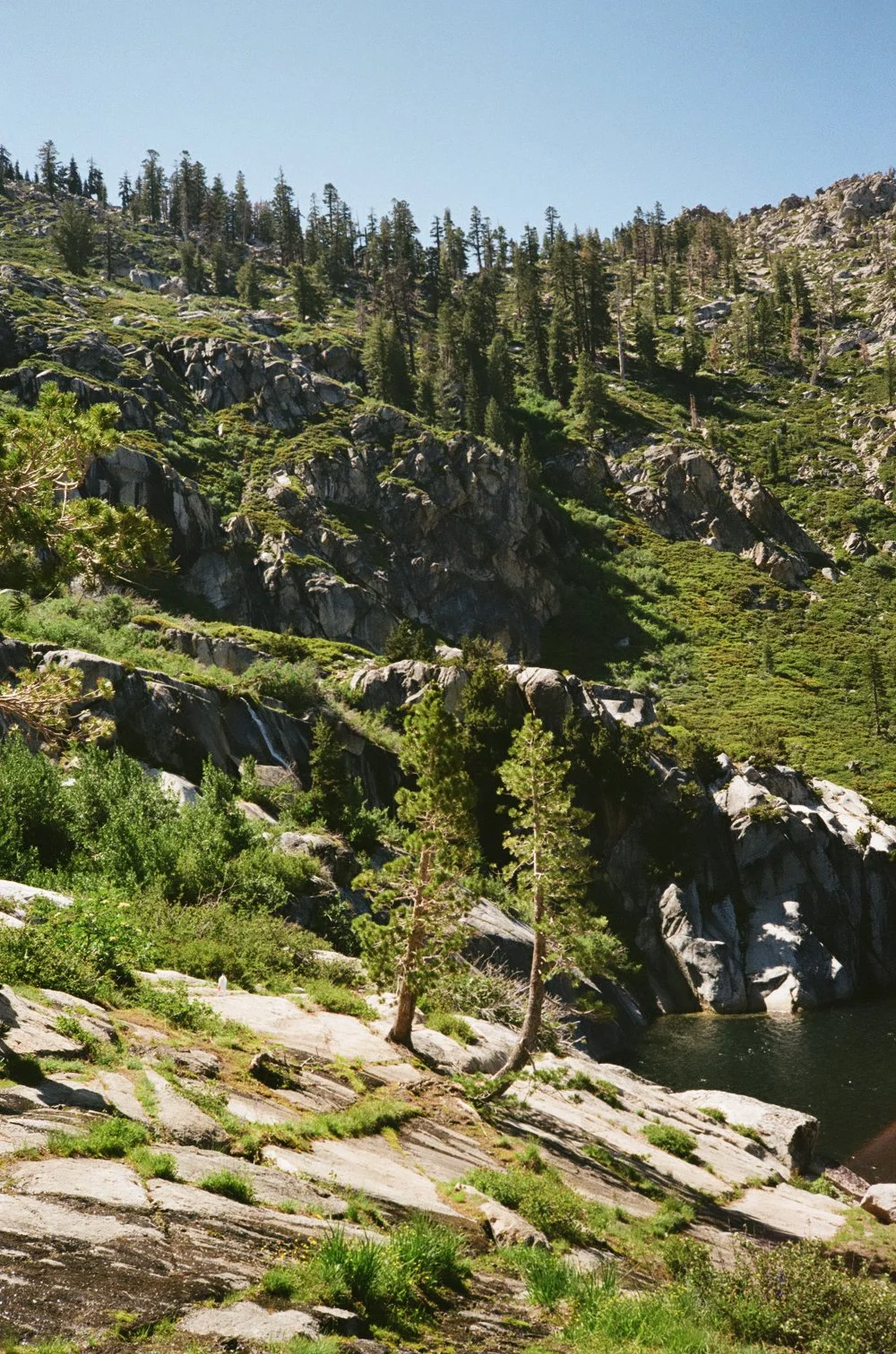 A mountainous landscape with rocky slopes, sparse trees, and a small body of water, under a clear blue sky.