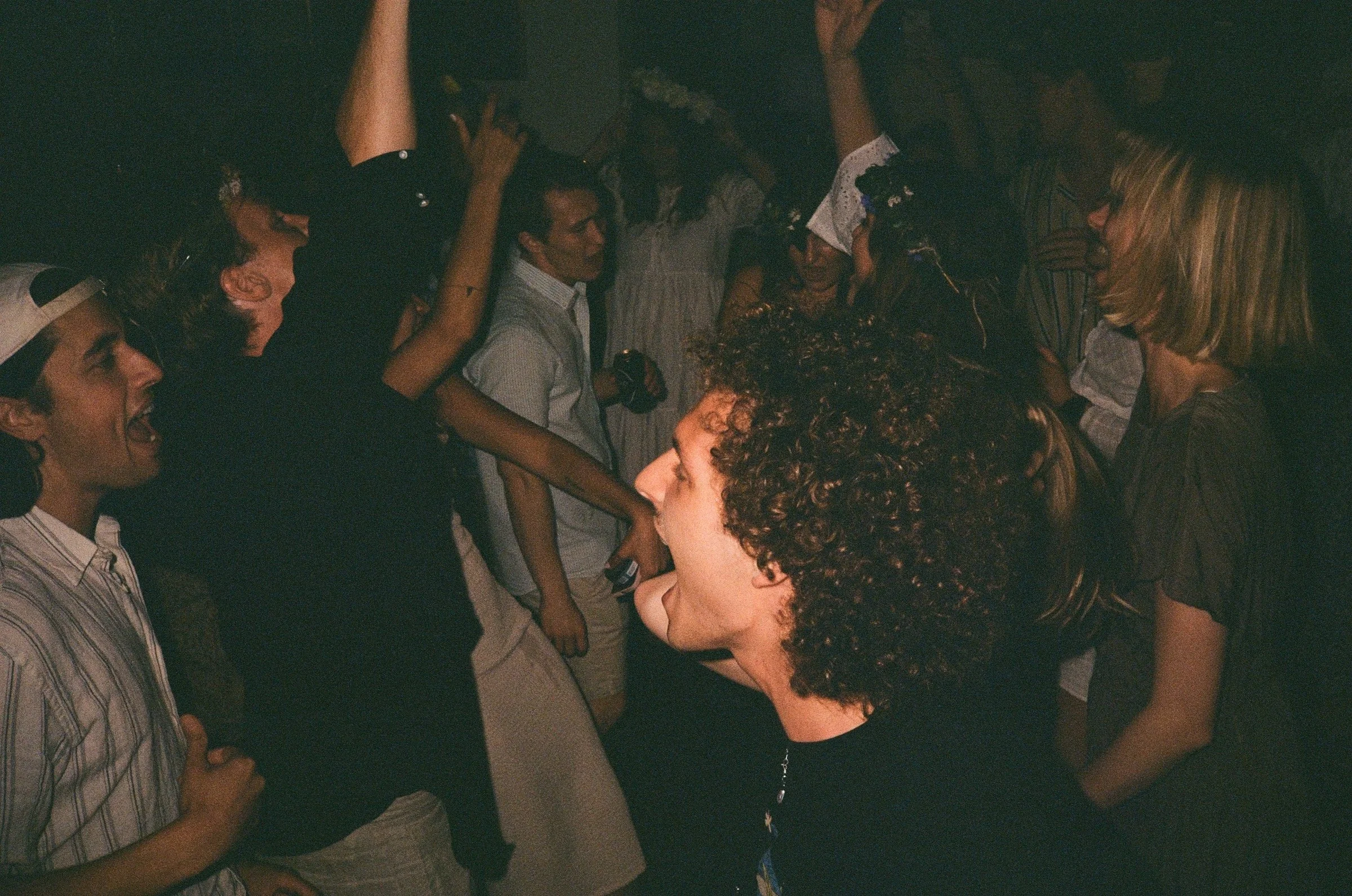 A group of people are standing closely together, celebrating around a cake with sparklers on top. They are smiling, laughing, and raising their hands in excitement.