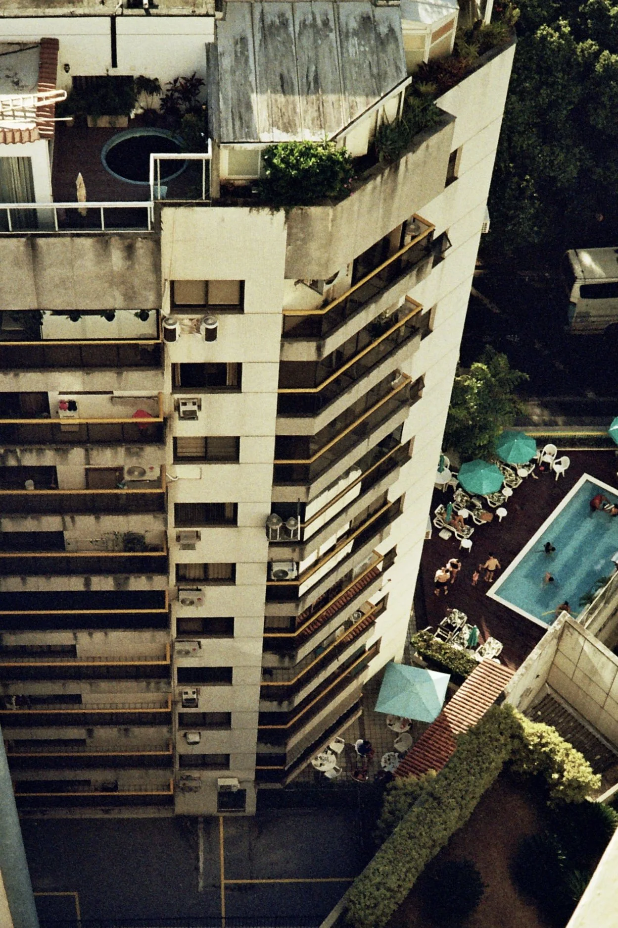 A view of a multi-story apartment building with balconies, a rooftop garden, a swimming pool, and a small outdoor seating area with umbrellas and people.