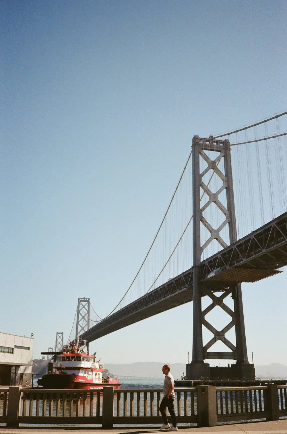 A bridge over water with a red boat passing underneath, and a man walking along the waterfront in the foreground.