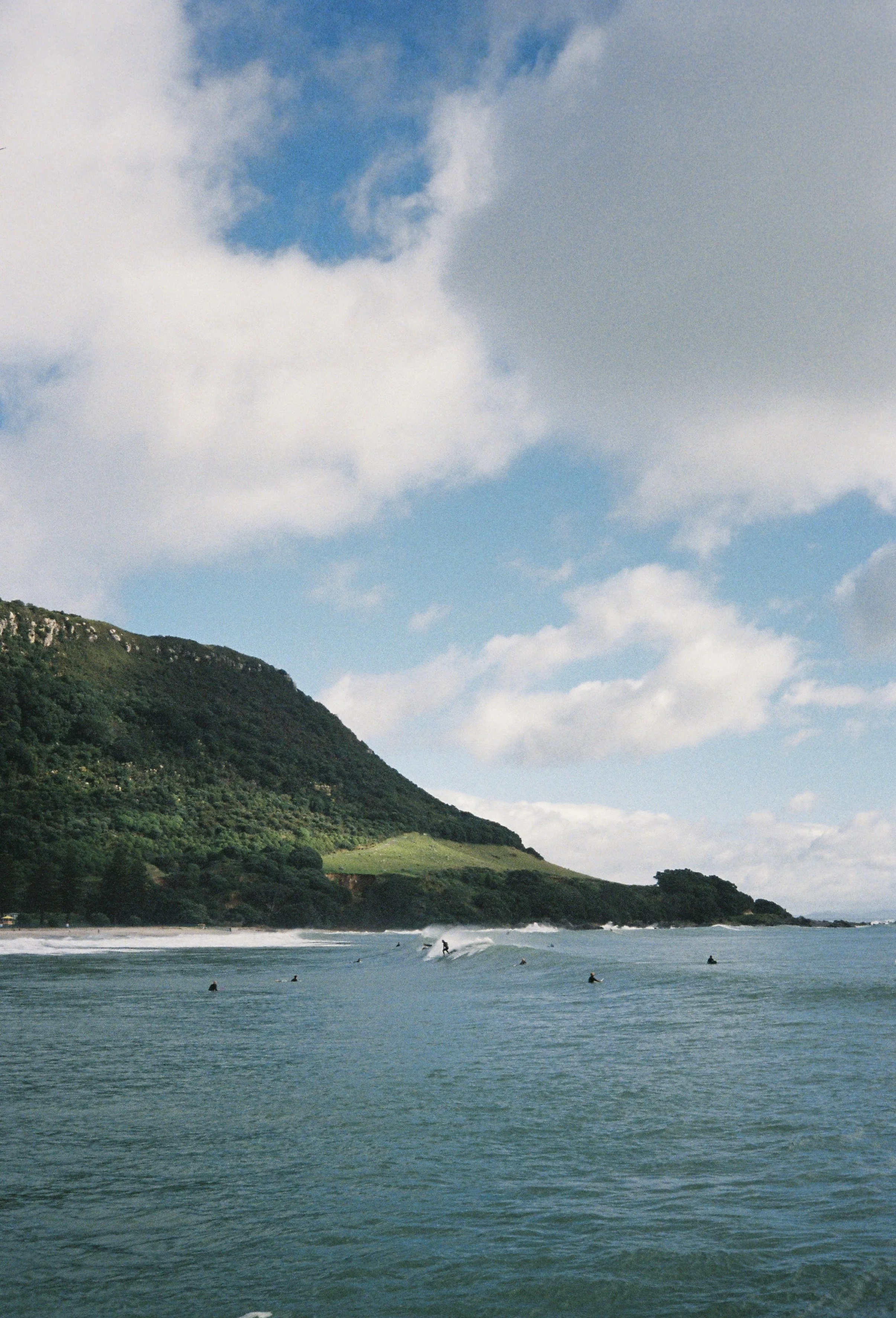 Ocean with surfers riding waves near a green hillside with trees and a cloudy sky.