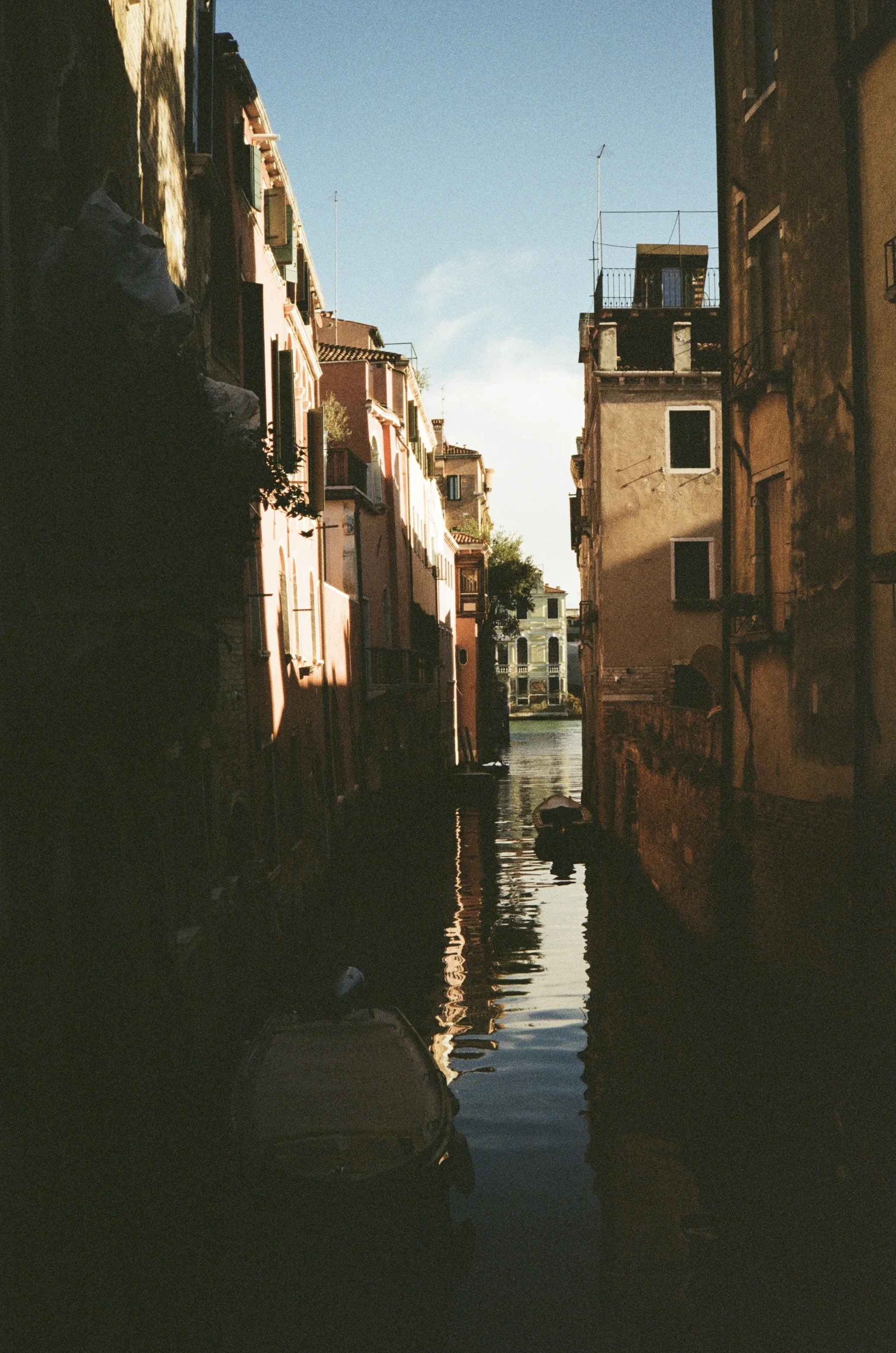 A narrow canal lined with old buildings on both sides, with boats moored along the edges. The water reflects the buildings and the sky, and there is a bright blue sky above.