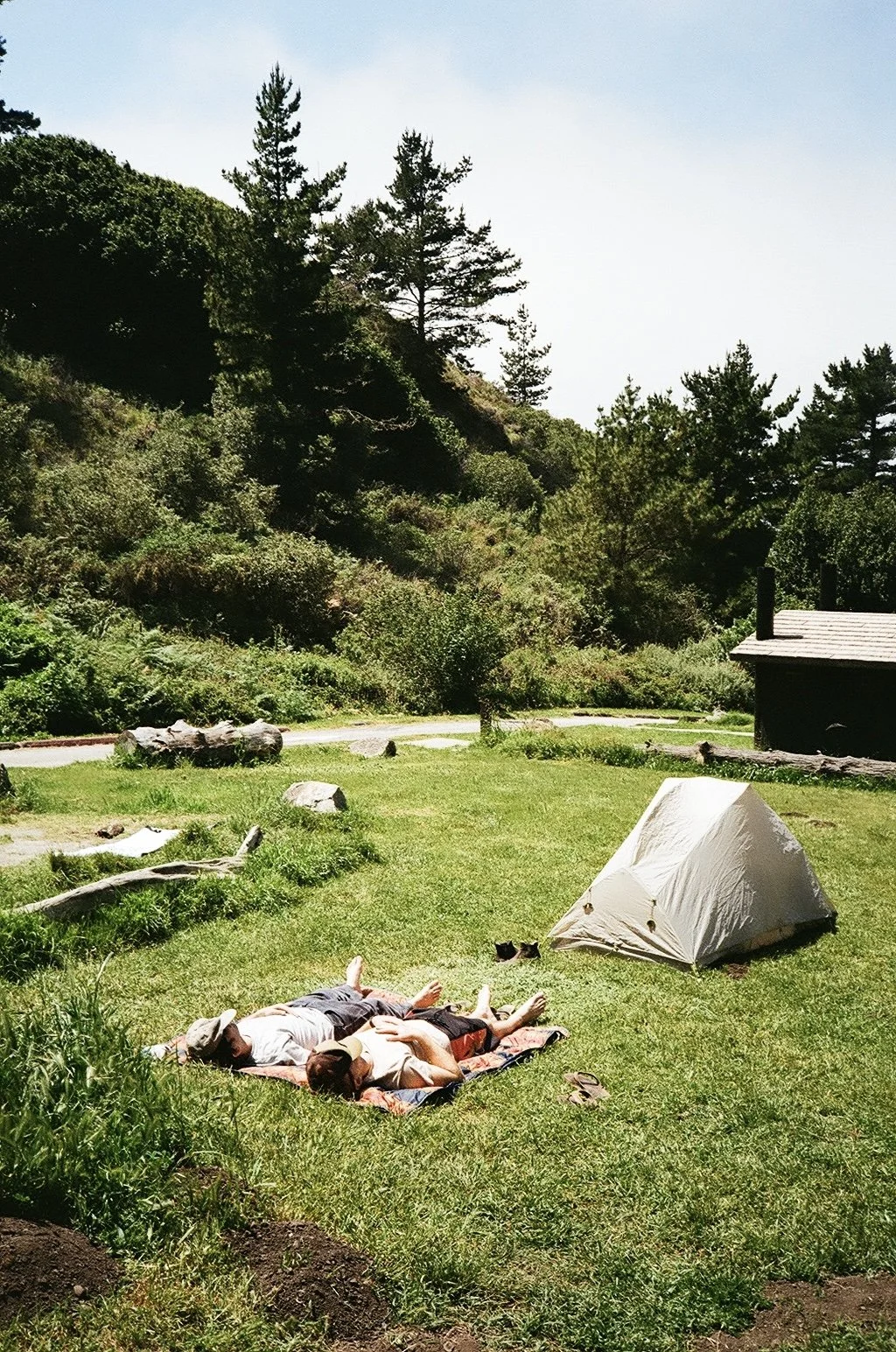 Two people lying on a blanket in a grassy campsite, with a small tent nearby, surrounded by trees and a wooden cabin in the background.