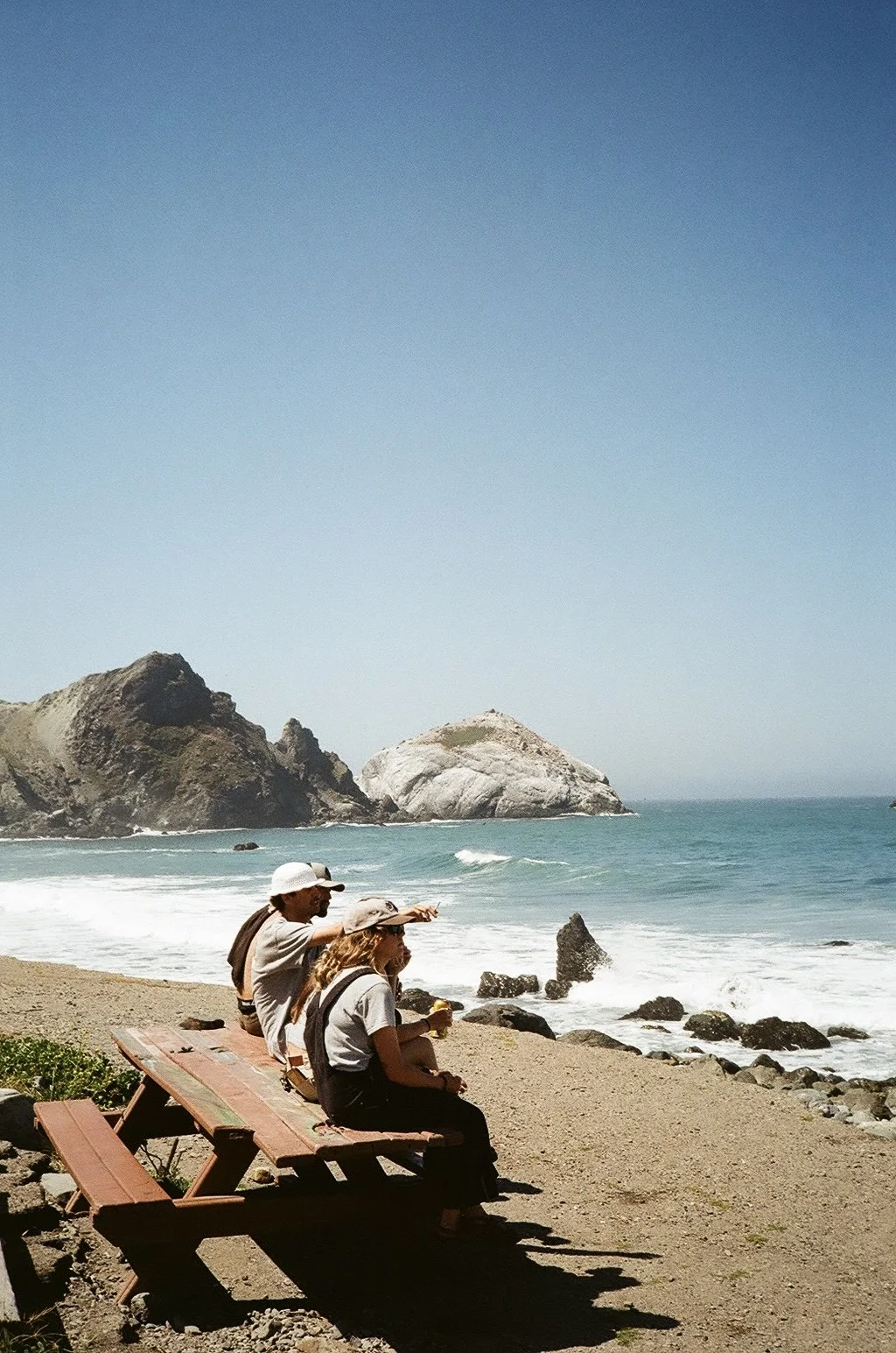 Three people sitting on a picnic table on a sandy beach, looking out at the ocean with waves and large rocks, under a clear blue sky.