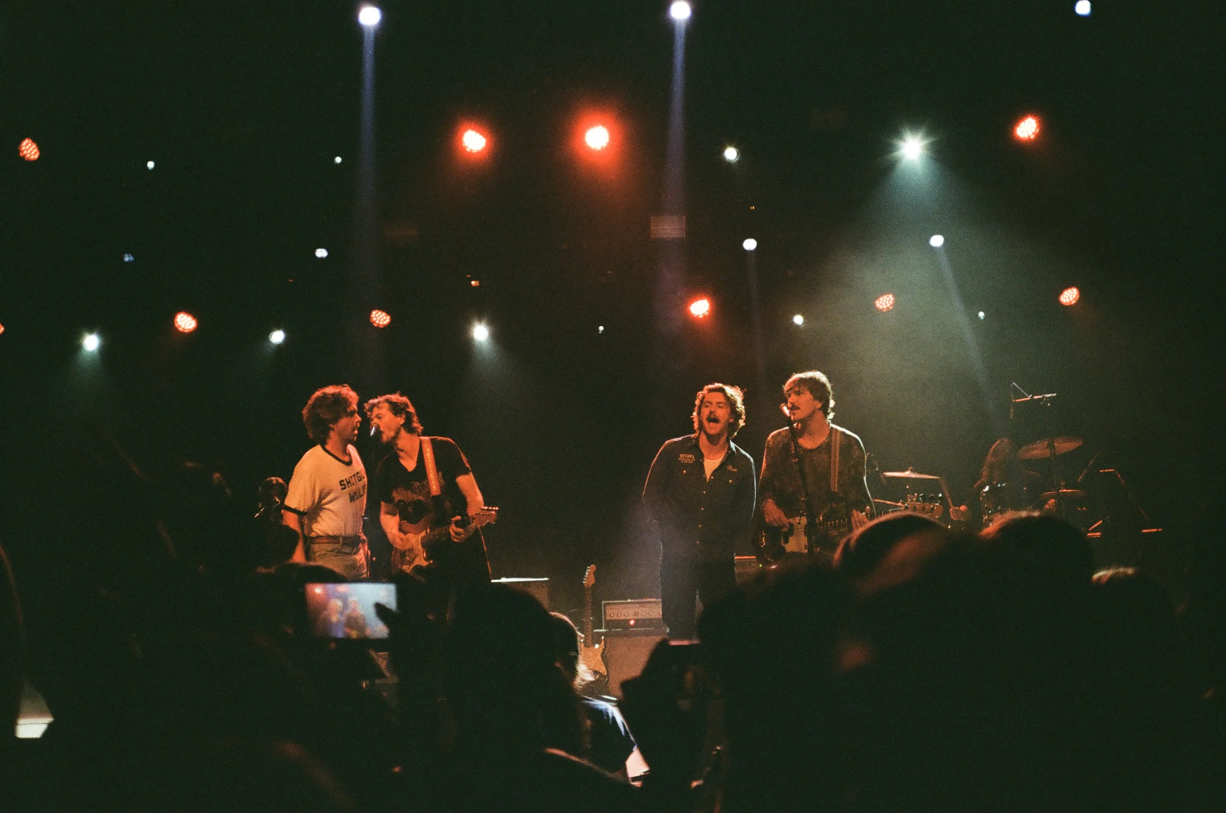 Musical band performing on stage with four members visible, illuminated by stage lights against a dark background.