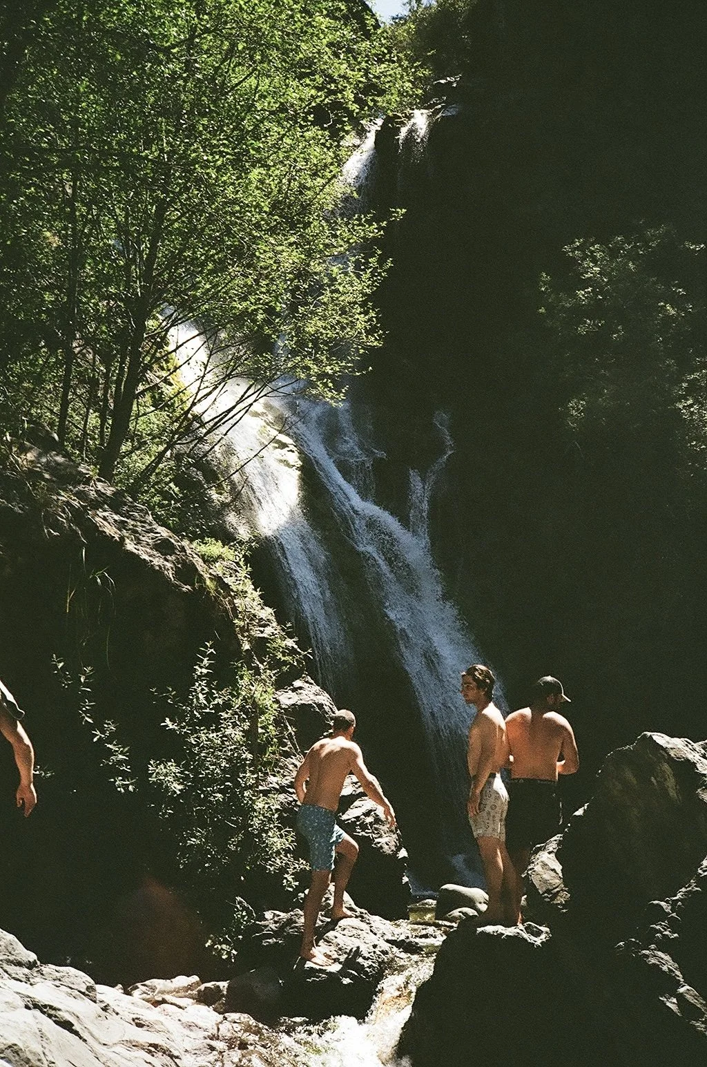 People swimming and relaxing in a rocky area near a waterfall surrounded by lush green trees.