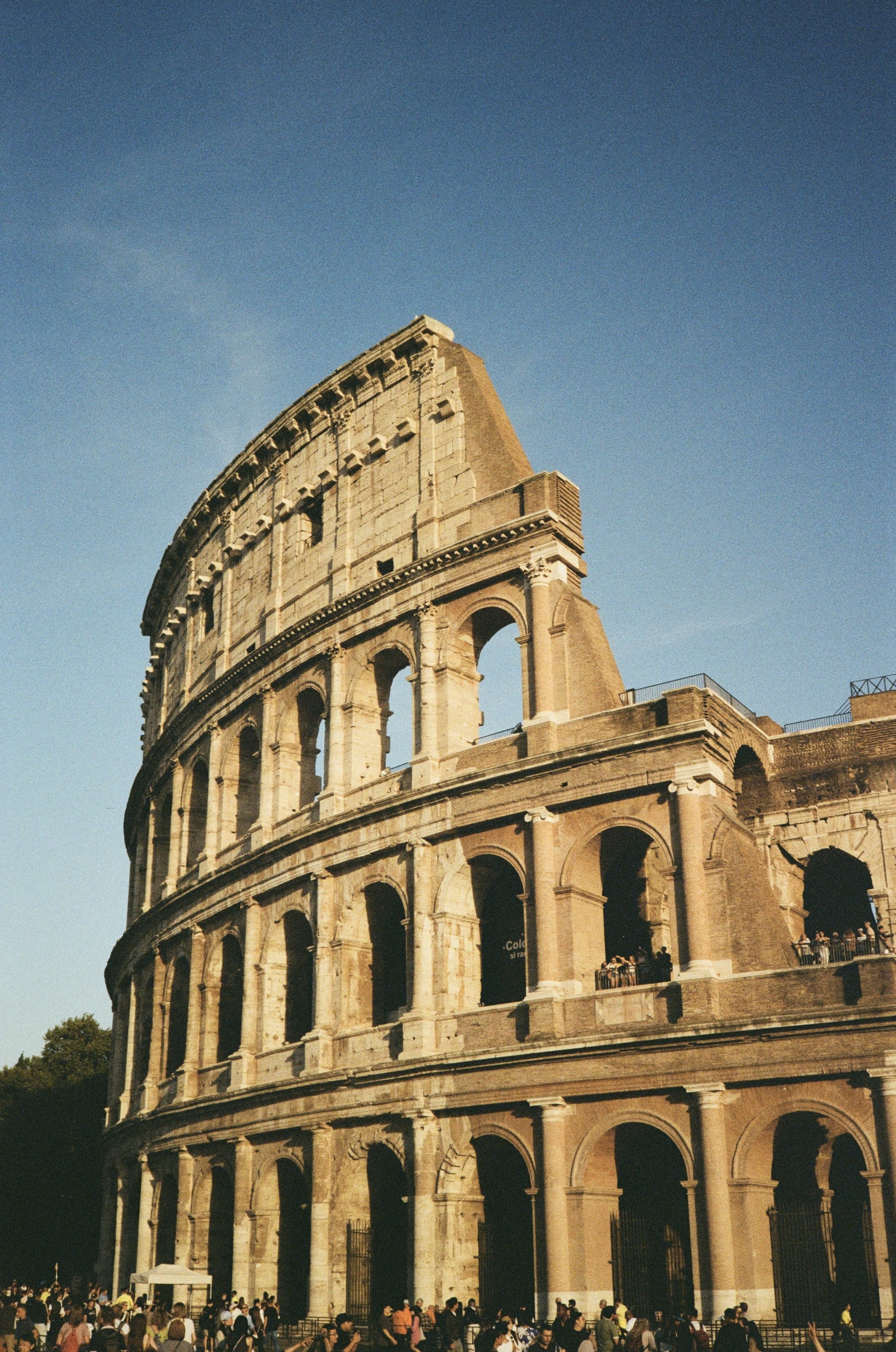 The image shows a partial view of the ancient Roman Colosseum with arches and tiers, and a large crowd of tourists gathered at the base against a clear blue sky.