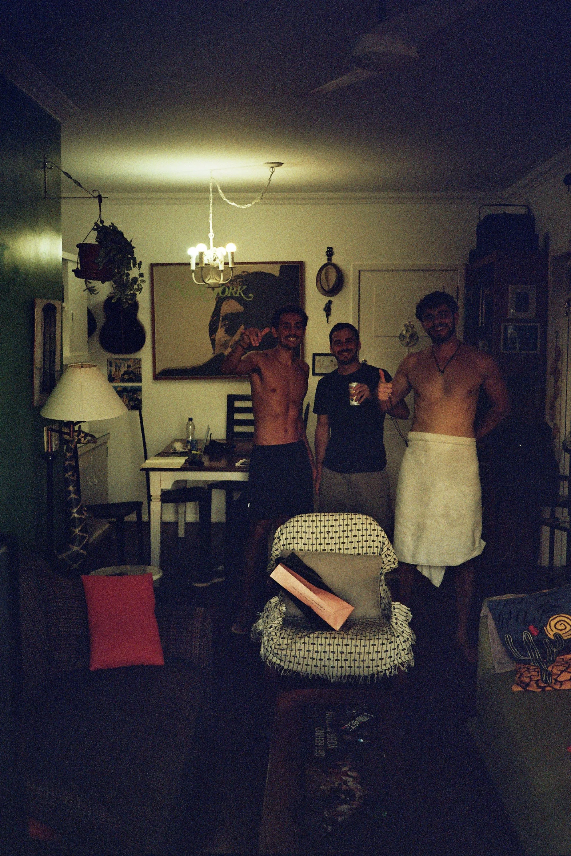 Three shirtless men standing indoors, one holding a can, all smiling, in a room decorated with artwork and musical instruments.