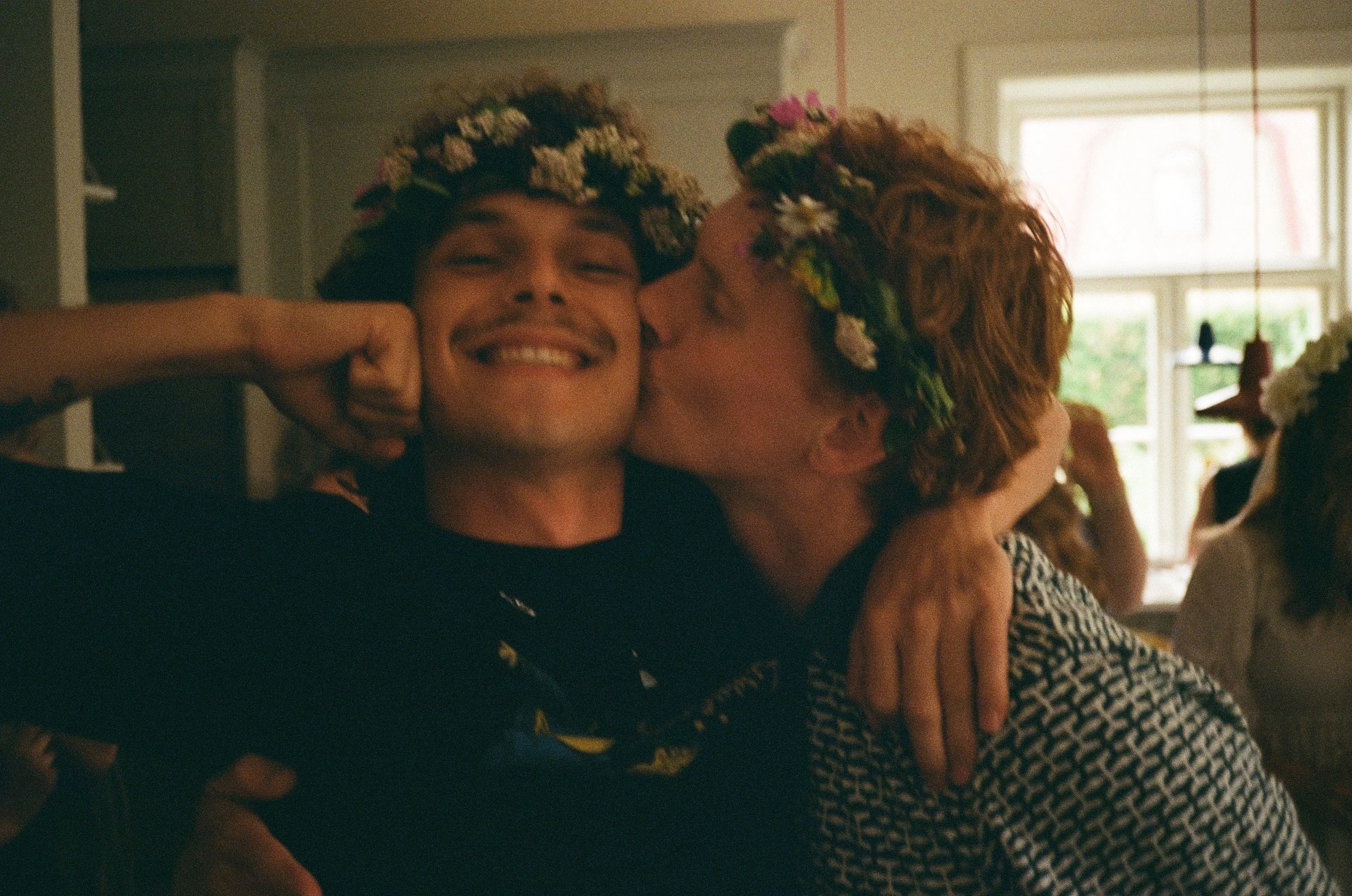 Two people embracing, wearing flower crowns, inside a room with large windows.