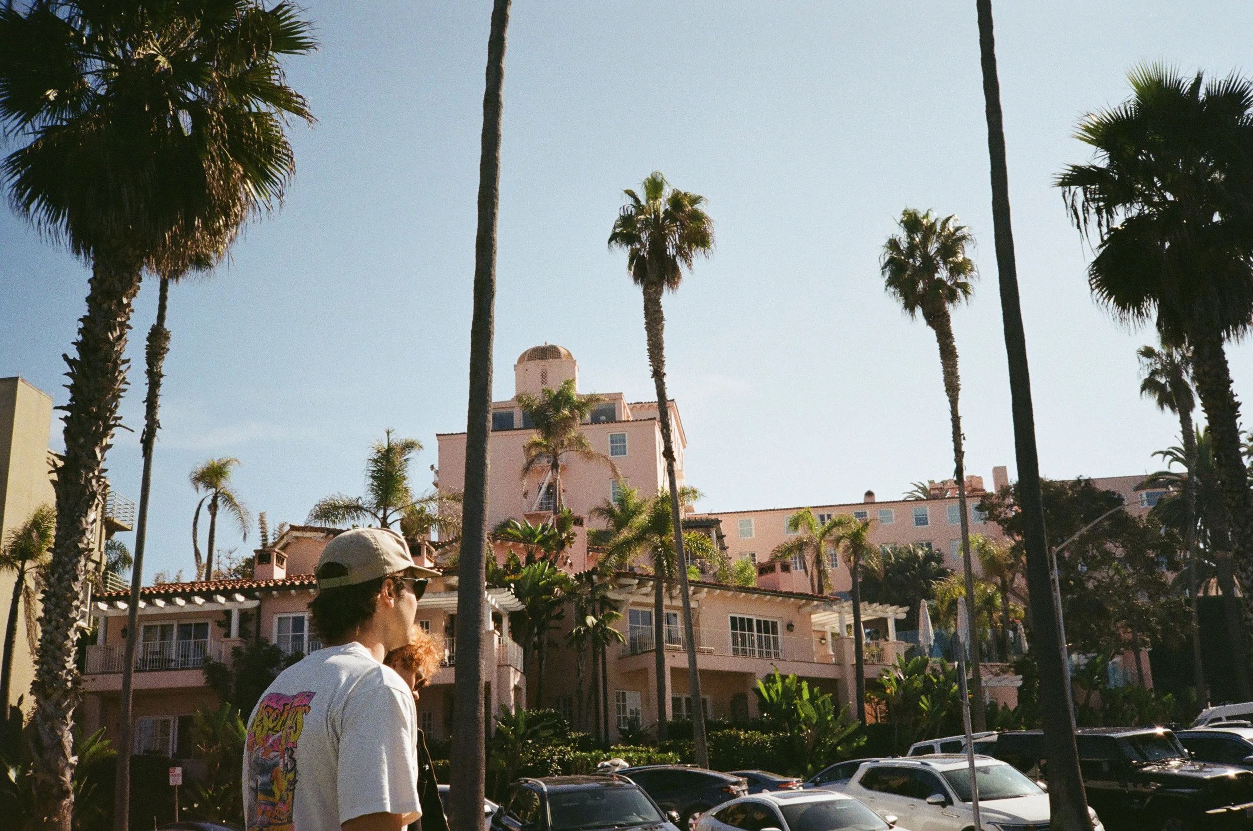 A sunny day in a city with palm trees and pink buildings, with a person in the foreground wearing a white cap and a graphic t-shirt.