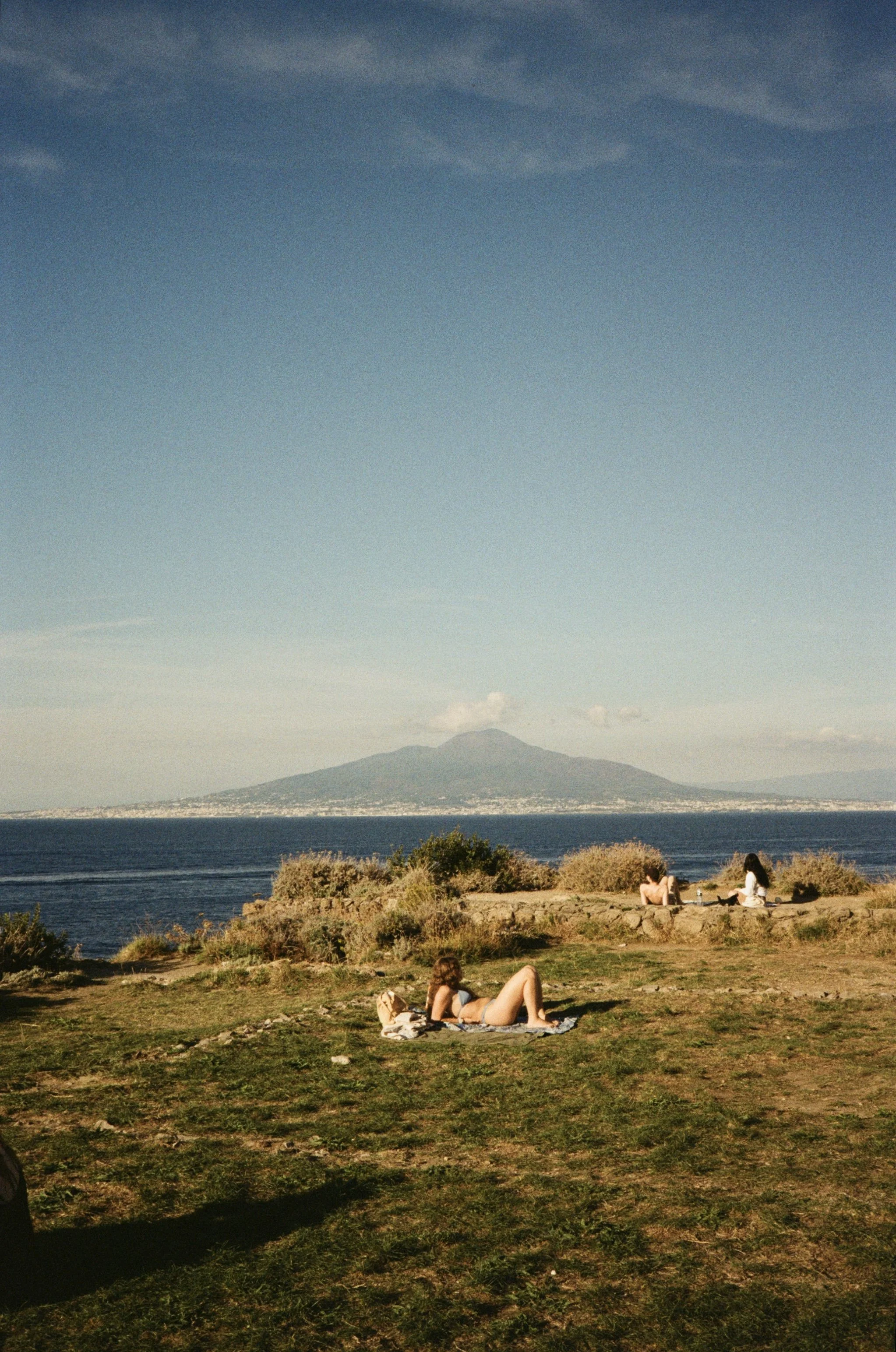 People relaxing on a grassy area near the coast, with a mountain in the distance and a blue sky above.