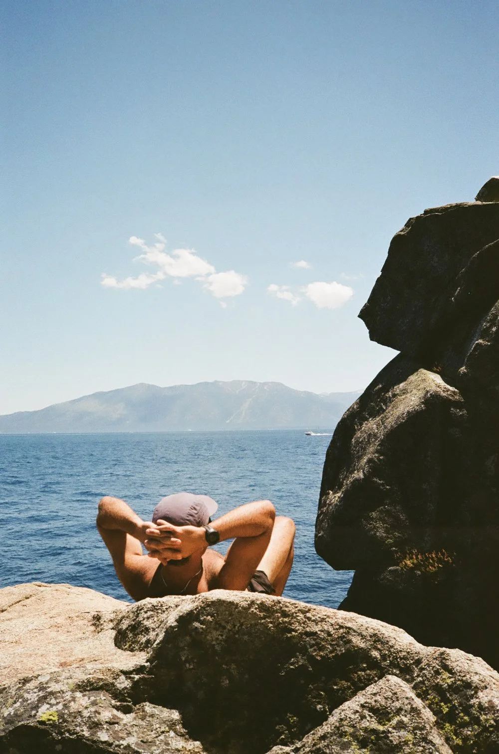 Person relaxing on rocks by the water with mountains in the distance under a blue sky with a few clouds.