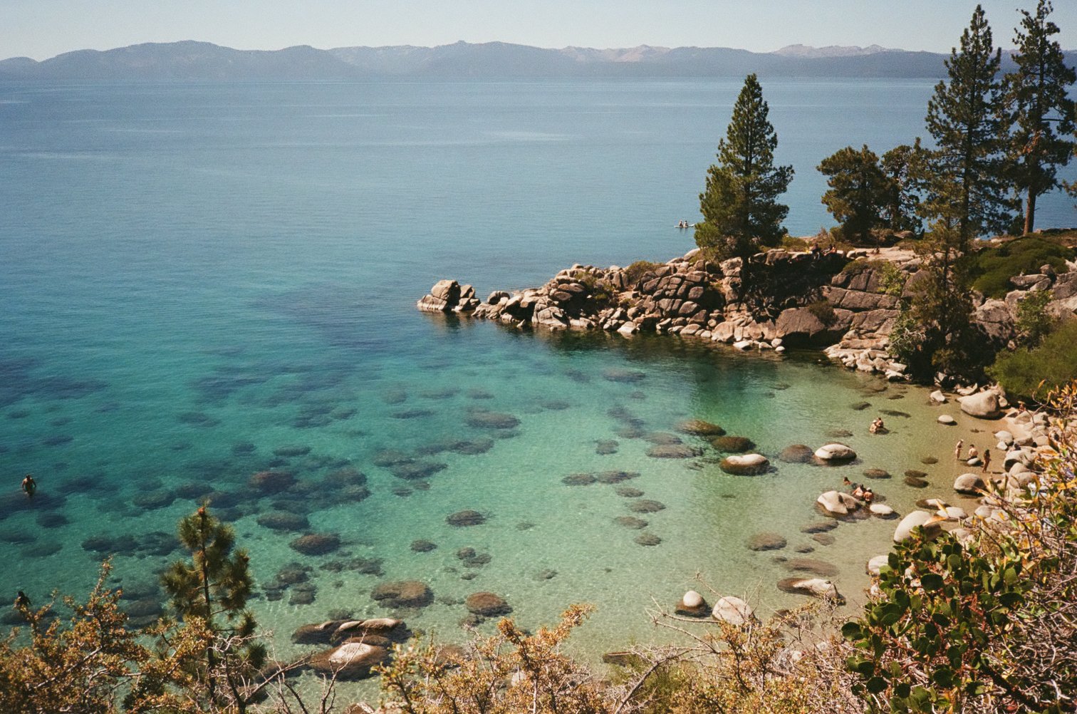 Scenic view of a rocky shoreline with clear turquoise water, pine trees on a small beach, and distant mountains in the background.