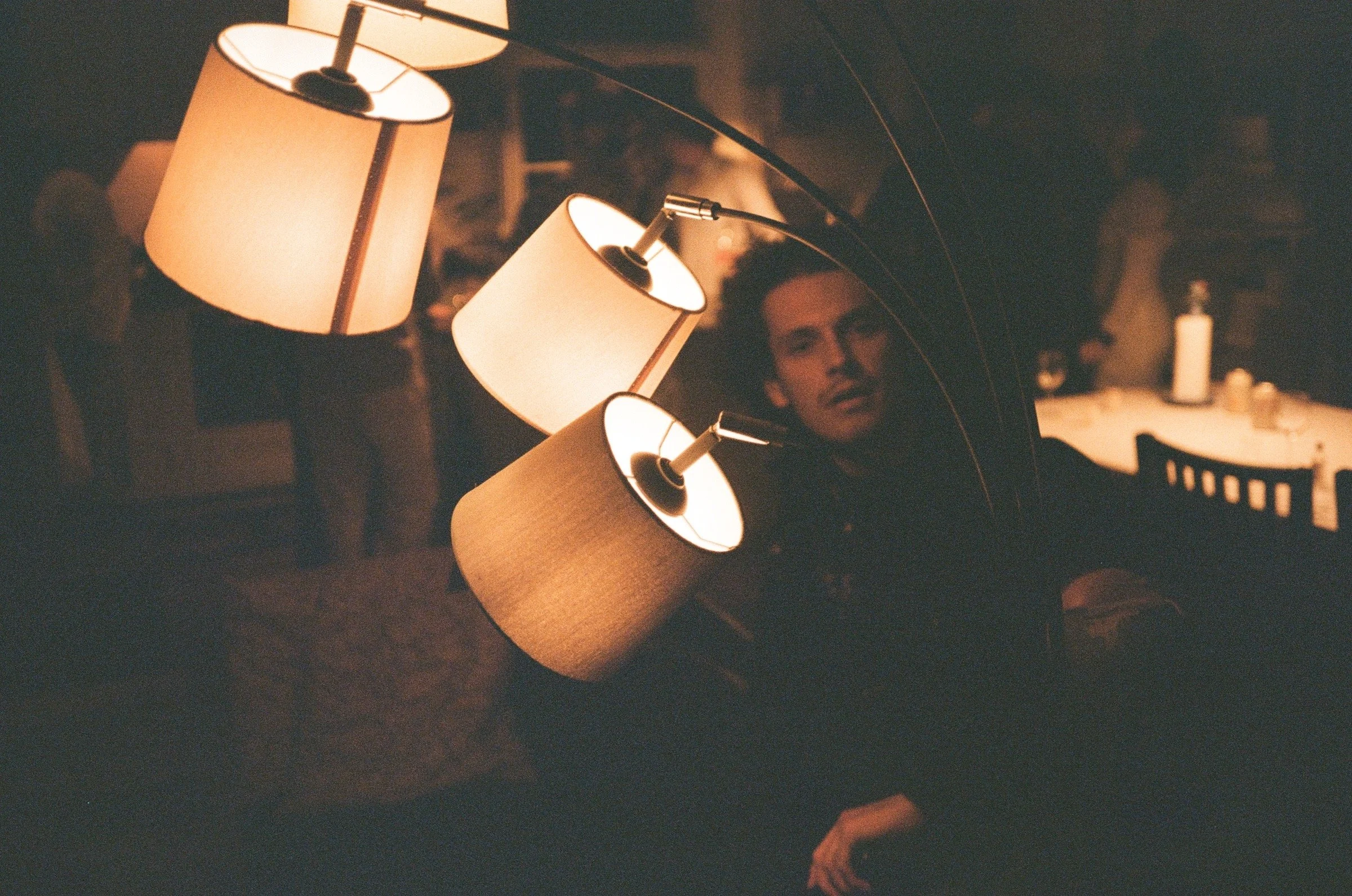 A person standing in a dimly lit room, partially obscured, with overhead lamps providing warm lighting, and a table with items in the background.