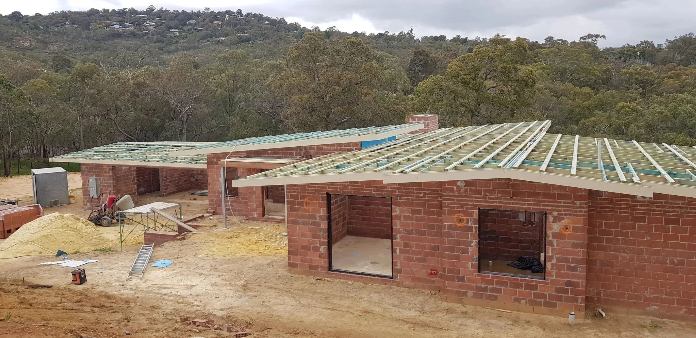 Construction site of a brick house with a partially built roof, surrounded by dirt and construction materials, with trees and hills in the background.