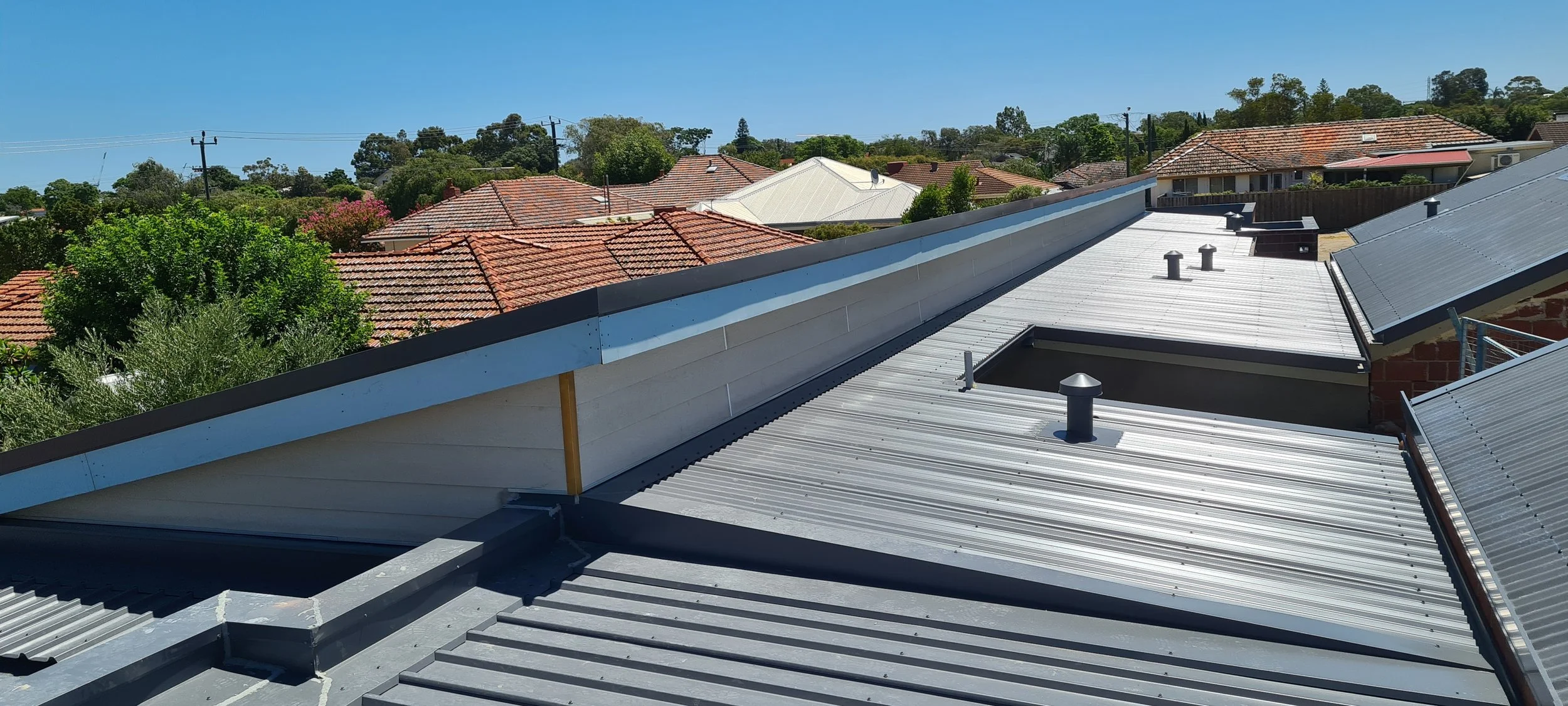 Several rooftops with metal and tile roofing under a clear blue sky, and trees in the background.