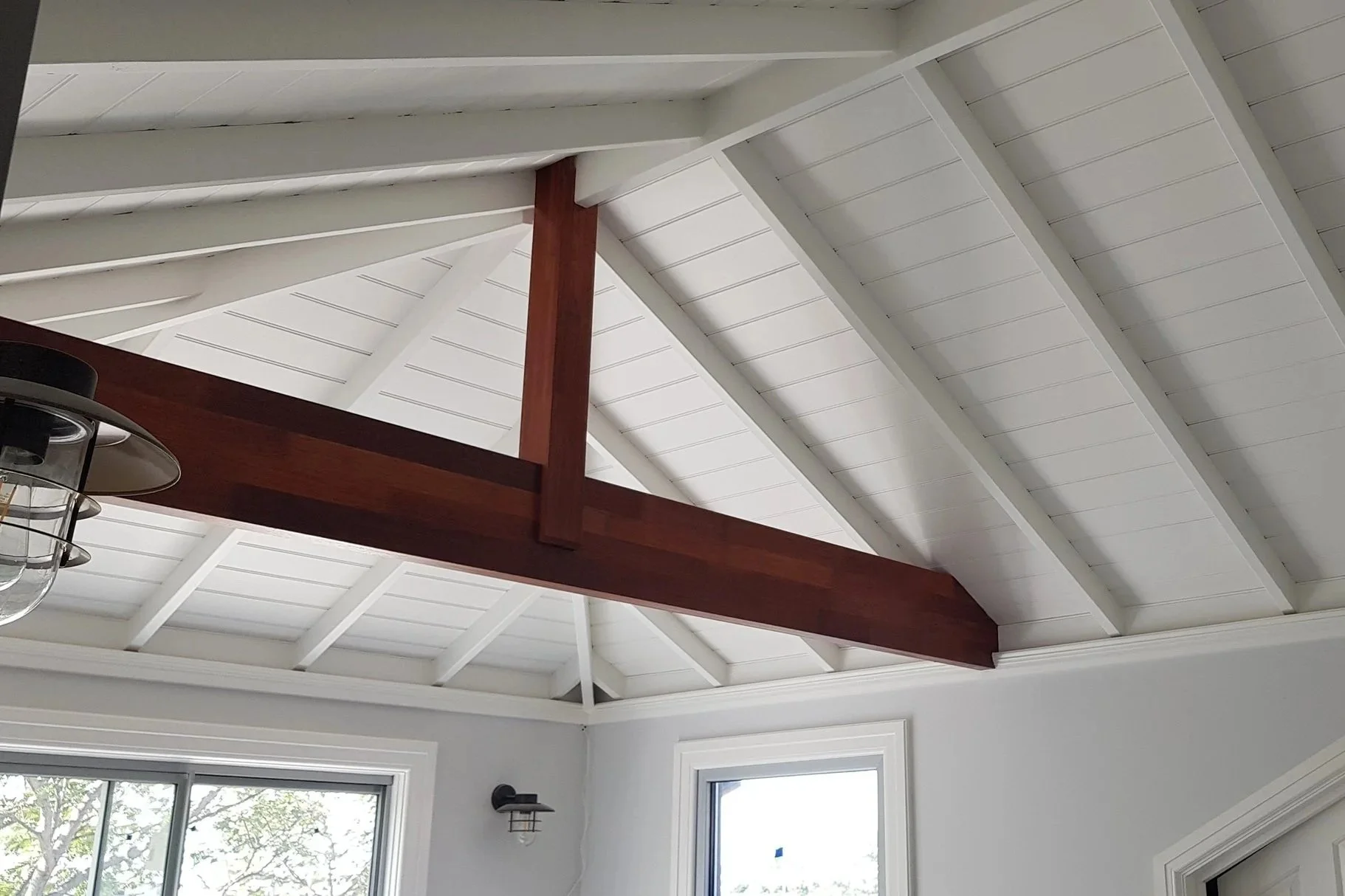 Interior view of a room with a white painted vaulted ceiling with exposed wood beams, large windows, and a wall sconce light fixture.