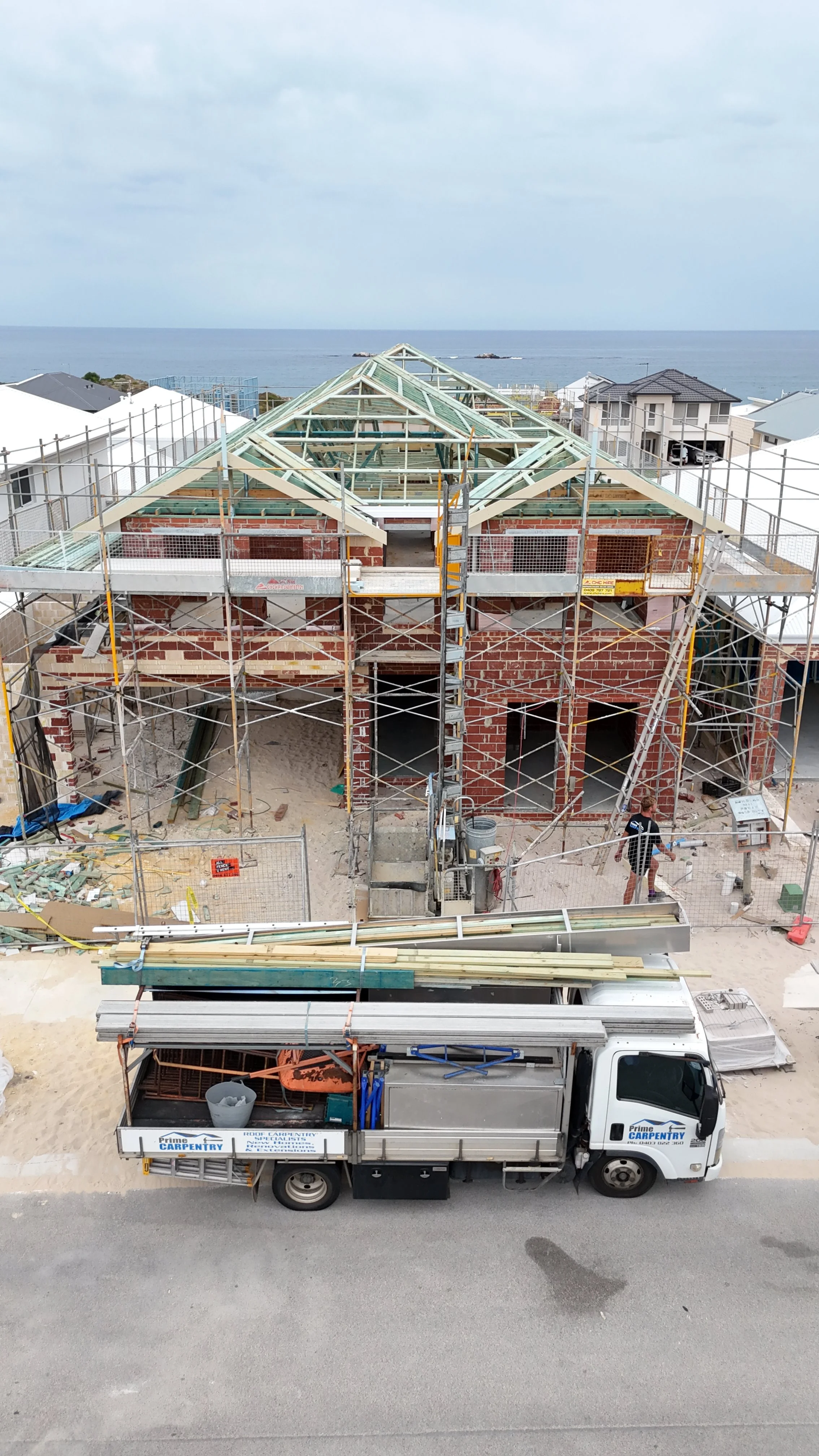 A house under construction near the beach with wooden framing and scaffolding, construction tools in a truck, and a worker on site.