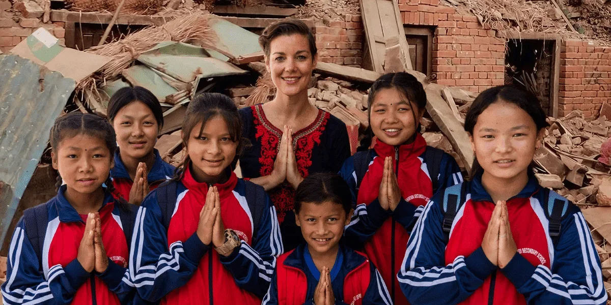 Group of seven young girls in sports uniforms with a woman standing behind them, all posing with hands together in a praying gesture outdoors amid debris and damaged buildings.