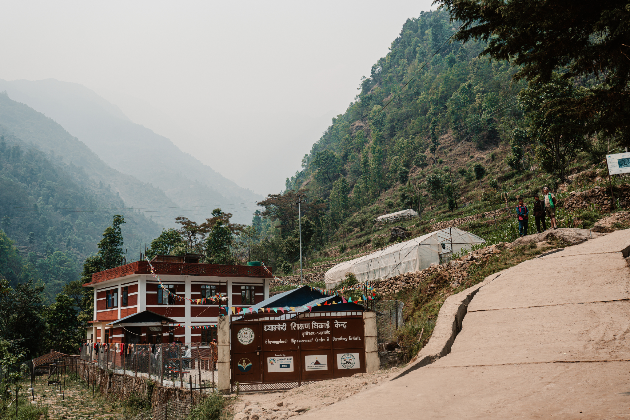 A mountainside scene with a two-story building, a gated entrance, and a sign that reads 'Ghyangephedi Improvement Centre & Dormitory for Girls.' There are three people standing on the roadside, and greenhouses or plastic tunnels on the hillside. The background shows forested mountains shrouded in haze.