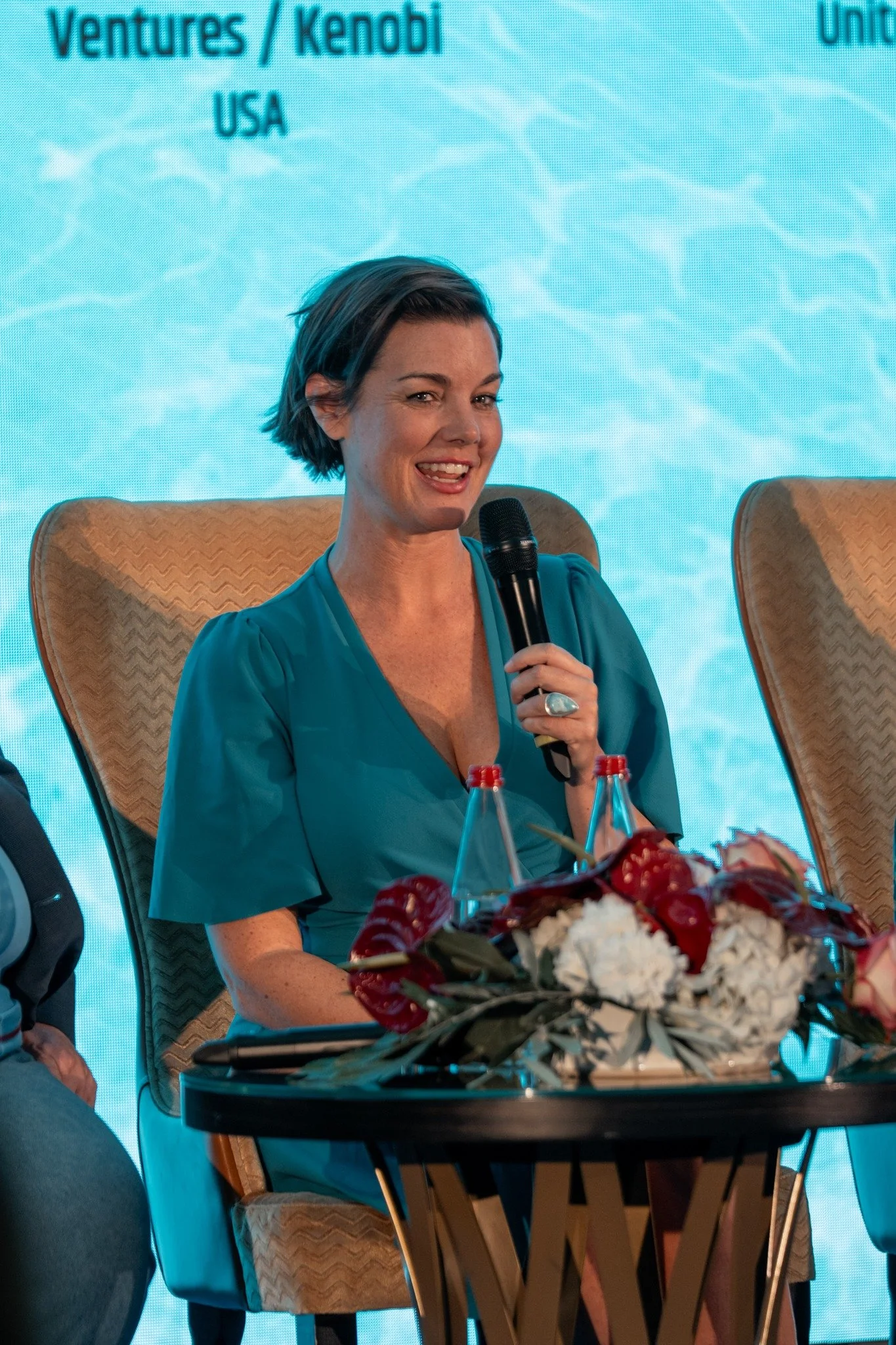 A woman holding a microphone speaking at a panel event, seated at a table decorated with flowers and bottles of water, with a blue background that has text related to venture capital and Kenobi.
