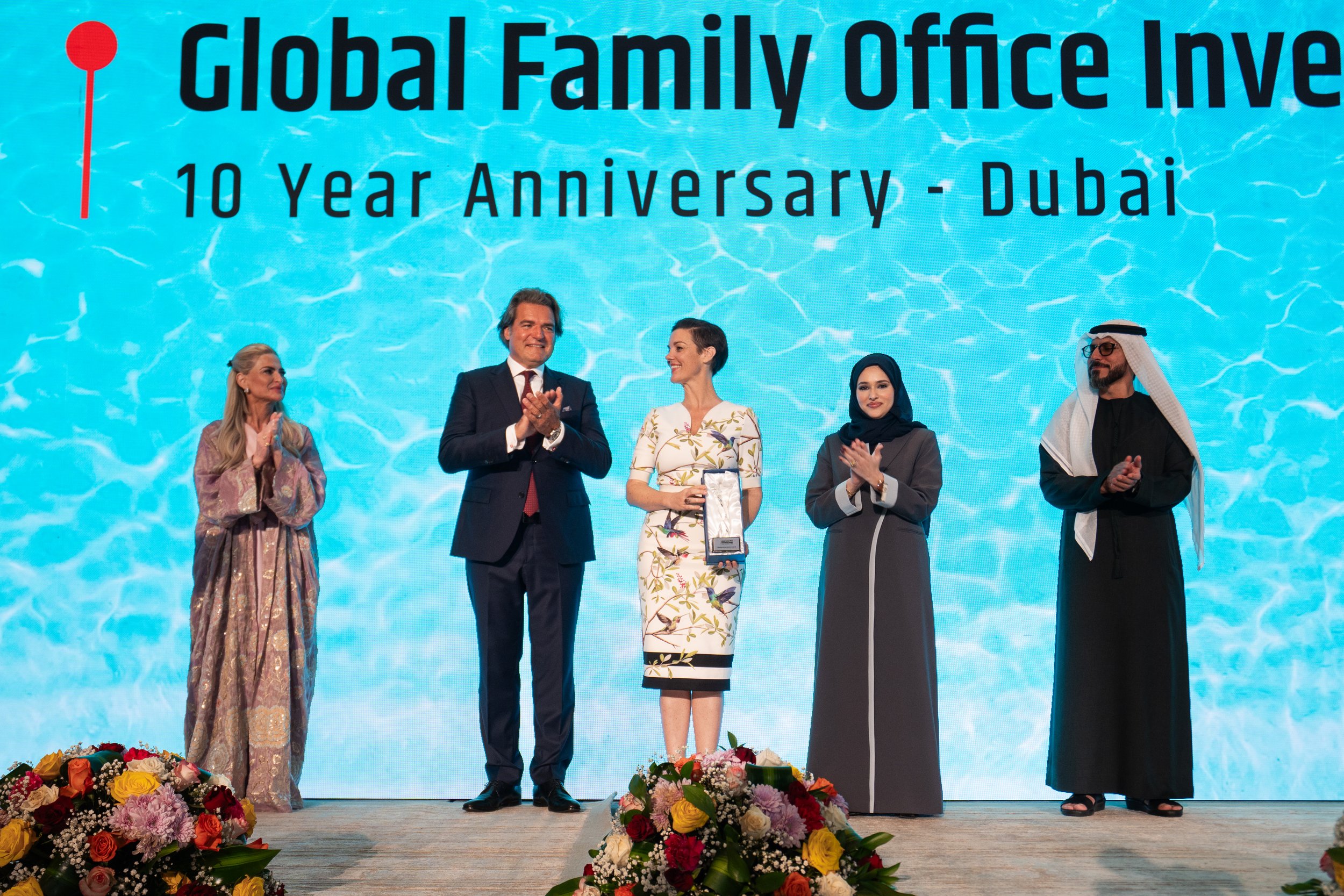 Group of five people on stage at a 10-year anniversary celebration for Global Family Office in Dubai. One woman is holding a trophy, while floral arrangements are in front of the stage, and a large screen with the event name is in the background.