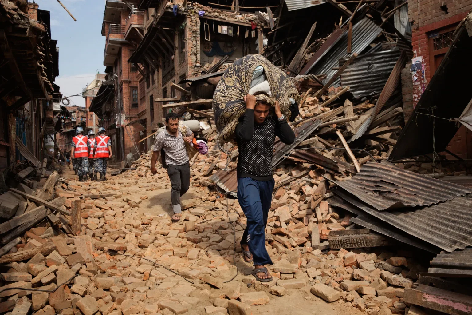 People walking through rubble and debris from collapsed buildings after an earthquake or natural disaster, with some wearing uniforms and helmets in the background.
