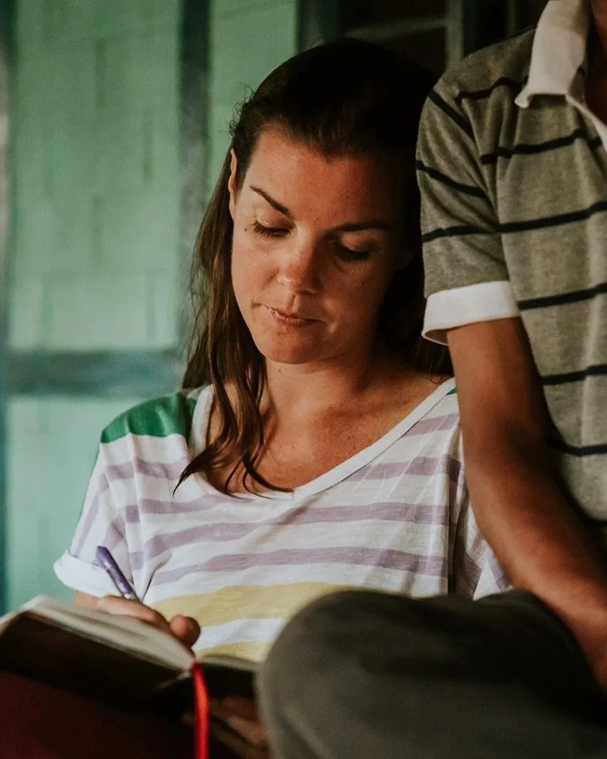 A young woman with long brown hair, wearing a white and pastel striped shirt, looking down at a book she is reading. A child's arm reaching over her shoulder, wearing a gray and black striped shirt. The setting appears to be indoors with green walls.