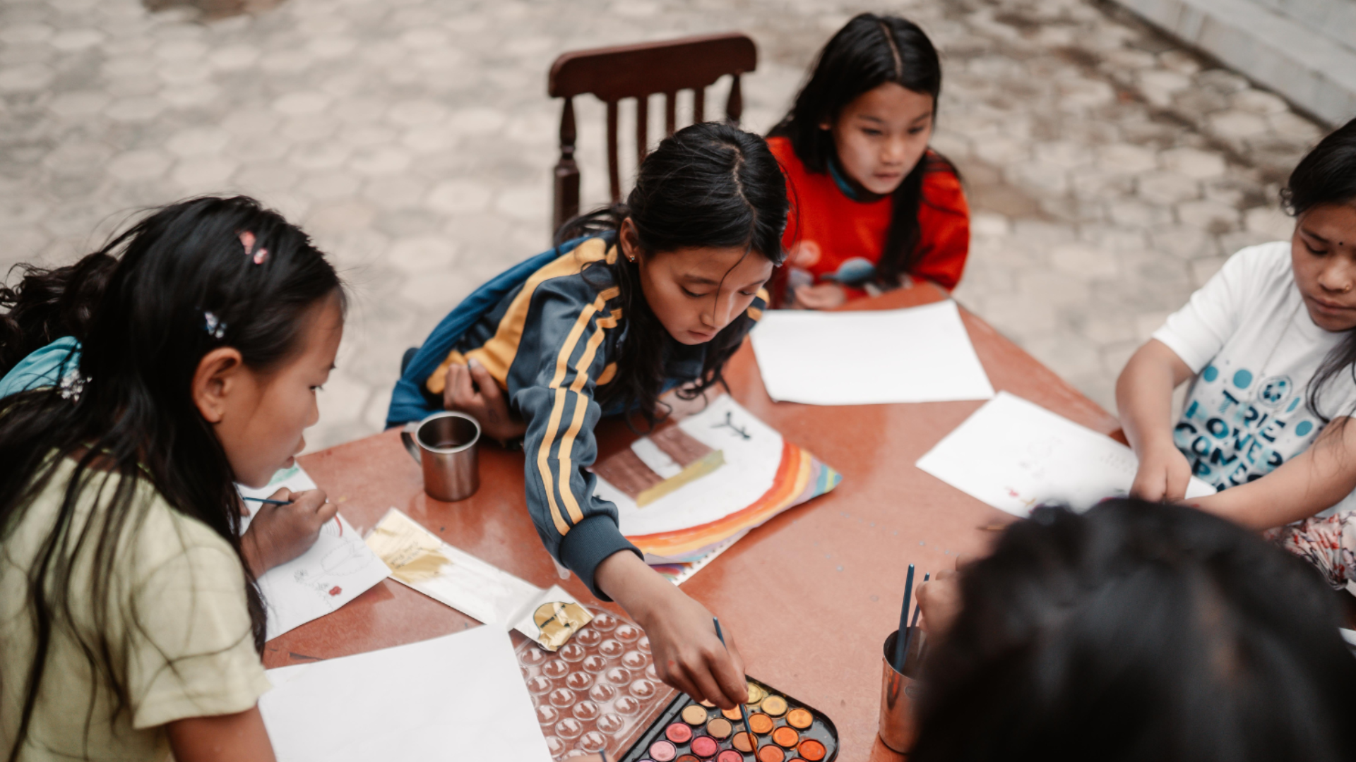 A group of children sitting around a wooden table, engaging in a painting activity with watercolor sets, papers, and art supplies outdoors.