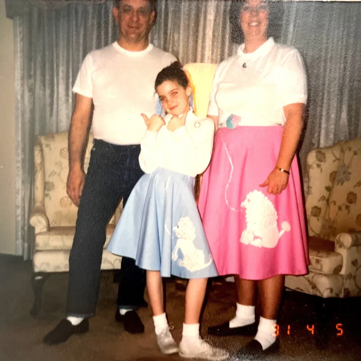A young girl standing between a man and a woman, all smiling at the camera. The girl is wearing a light blue skirt and a cream top, with her hand on her face and making a playful gesture. The man is in a white T-shirt and dark pants, and the woman is in a white polo shirt and a pink skirt with a white dog design. They are indoors in a living room with floral patterned chairs and curtains.
