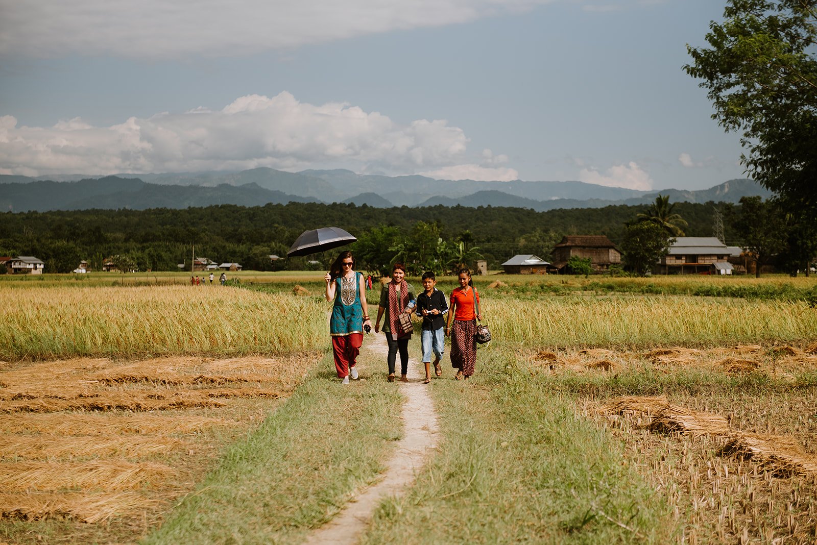 Group of five people walking along a narrow dirt path through a rice field under a cloudy sky, with mountains and houses in the background.