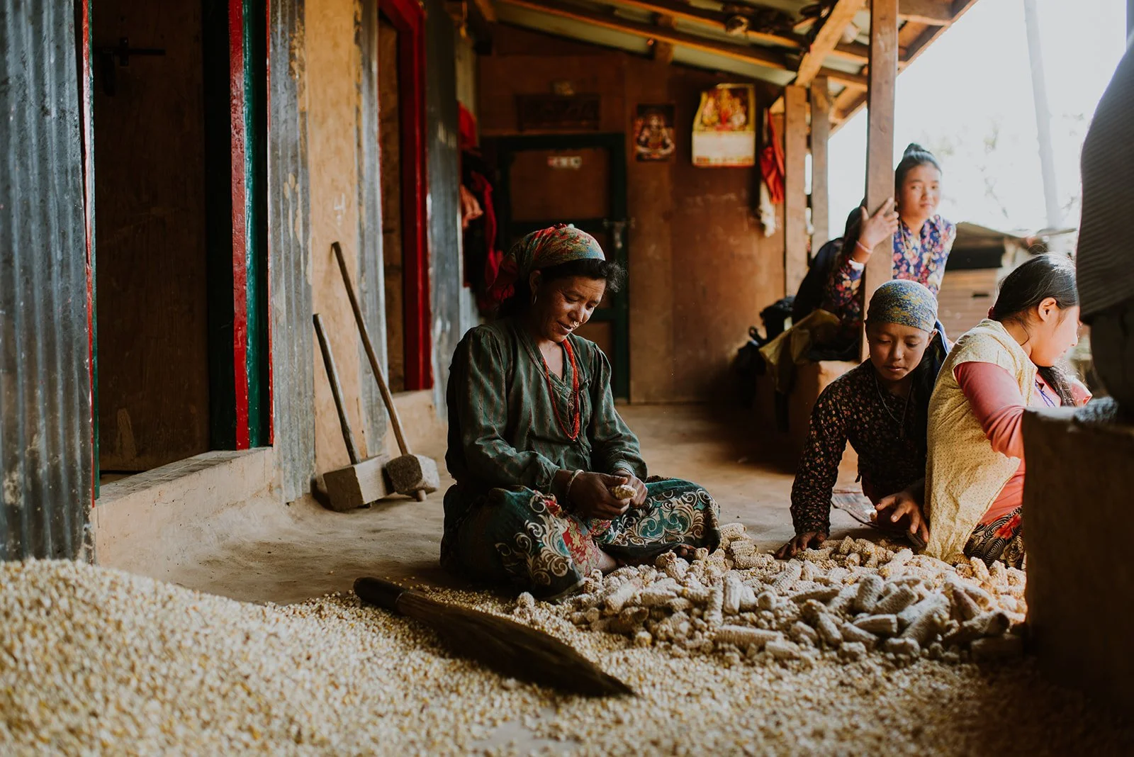 Women working with corn or grains inside a rustic room with wooden and metal walls, some women sitting and sorting grains, others standing nearby.