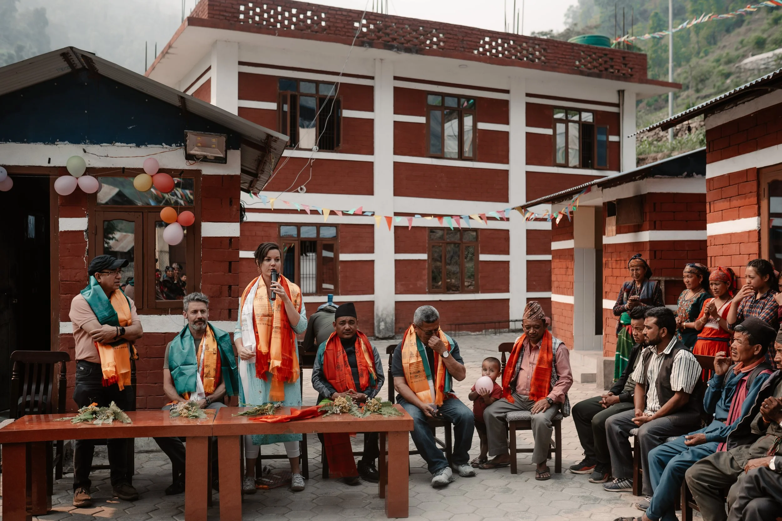 People gathered outdoors in front of red brick buildings for a cultural ceremony, with colorful decorations and floral arrangements on the table.