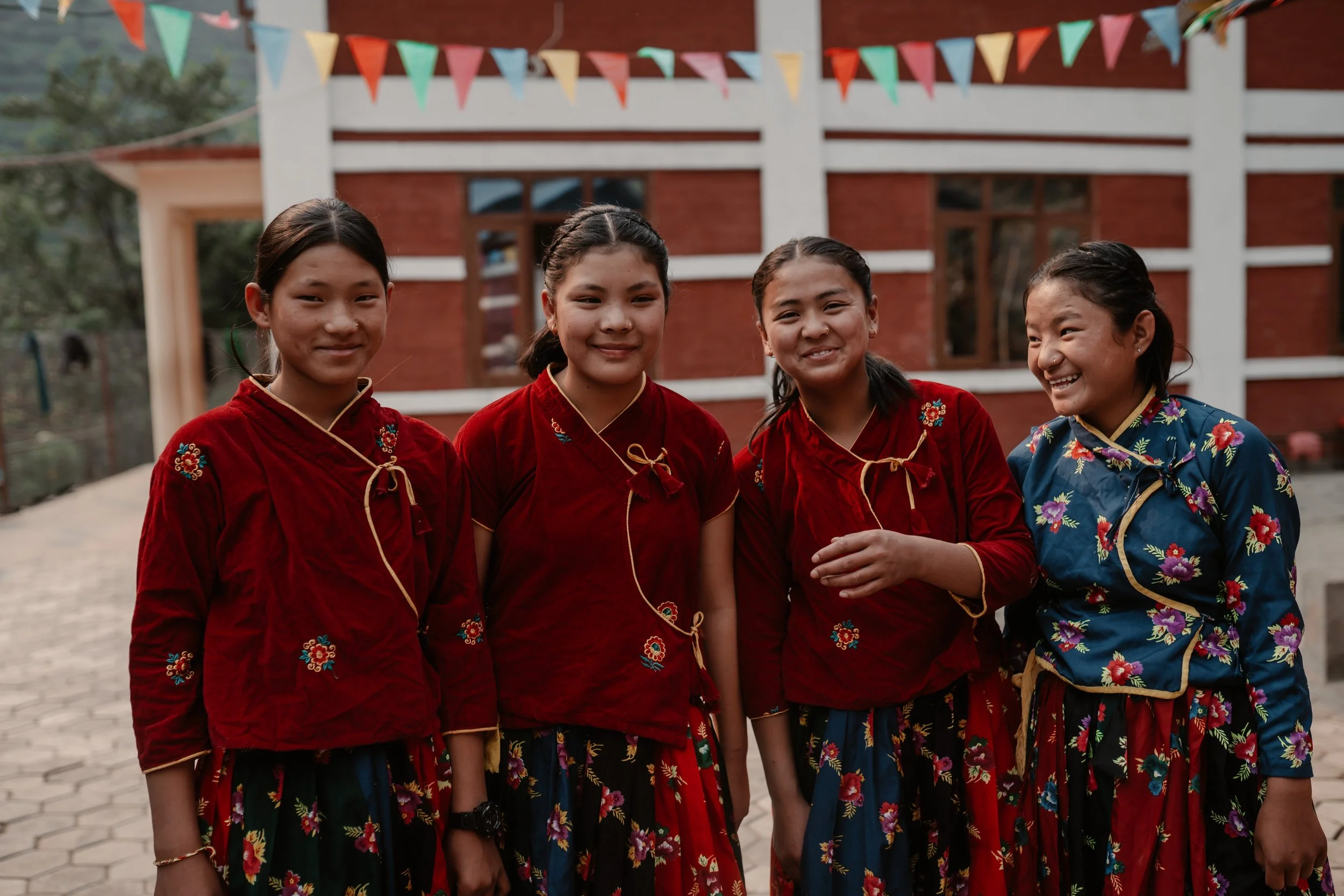 Four young girls standing together outside, dressed in traditional colorful clothes, smiling, with a brick building and colorful flags overhead in the background.