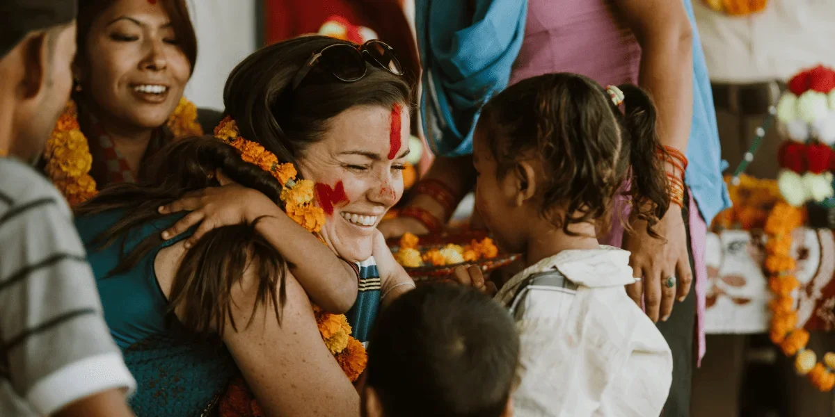 A woman with traditional face paint and garlands hugging and smiling at a young girl during a celebration with other smiling people around.